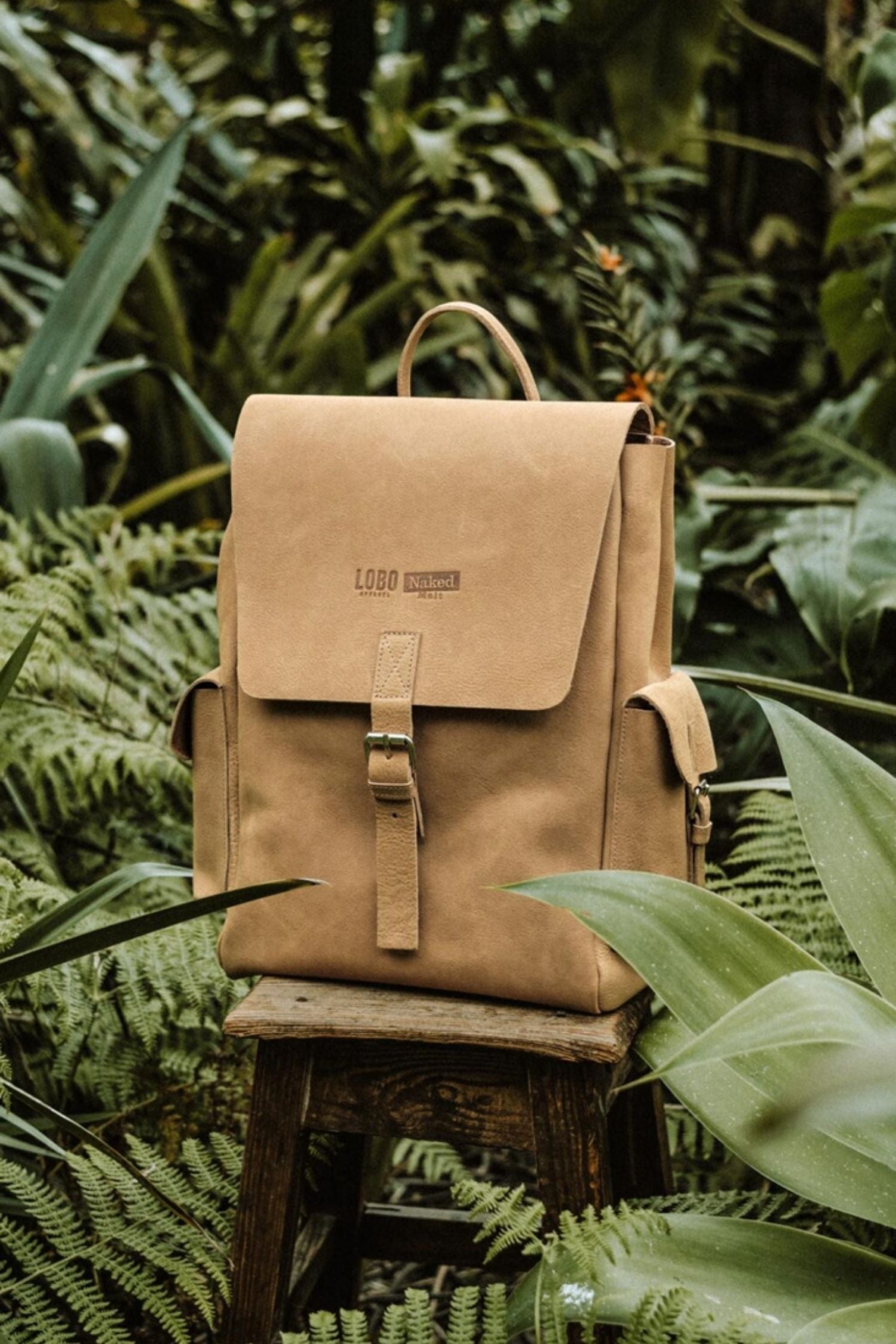 Lobo Apparel x Naked Malt brown leather backpack, minimalist design with center buckle, placed on a wooden stool in a natural outdoor setting.