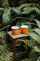 Pair of orange enamel mugs from the Lobo x Naked Malt collaboration on a rustic wooden stool in a natural outdoor setting.