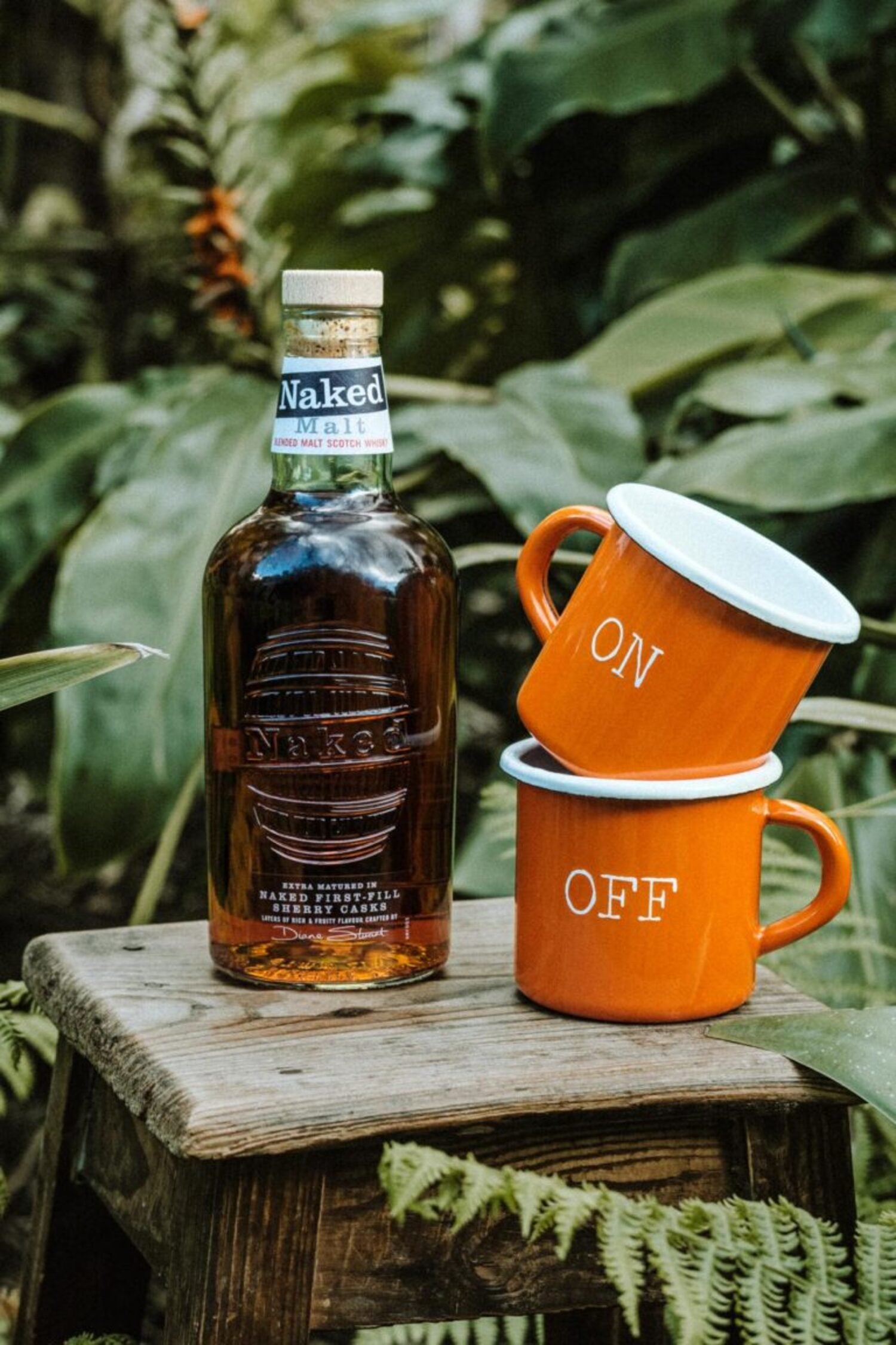 Lobo Apparel ON OFF mugs next to a Naked Malt whisky bottle in a tropical forest setting.