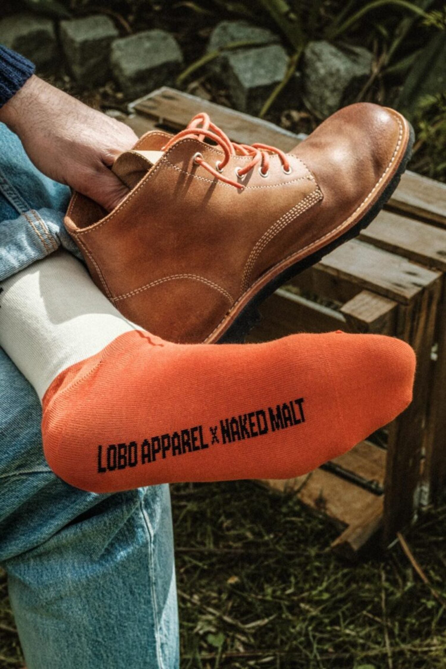 Man wearing Lobo leather boots with special edition orange socks from the Lobo x Naked Malt collection in a rugged outdoor setting.