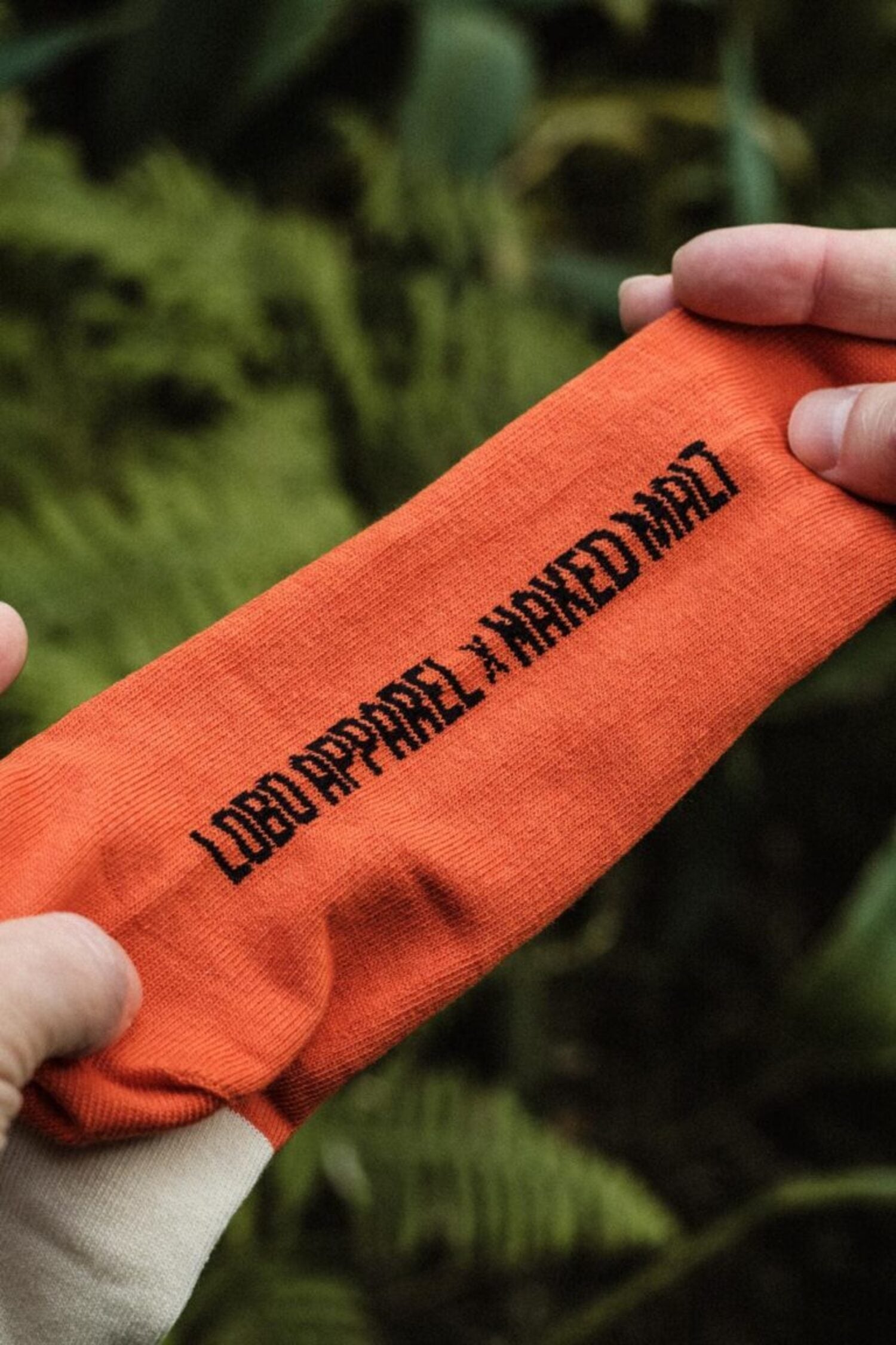 Hands stretching a vibrant orange Lobo Apparel x Naked Malt collaboration sock, highlighting the premium knit texture.