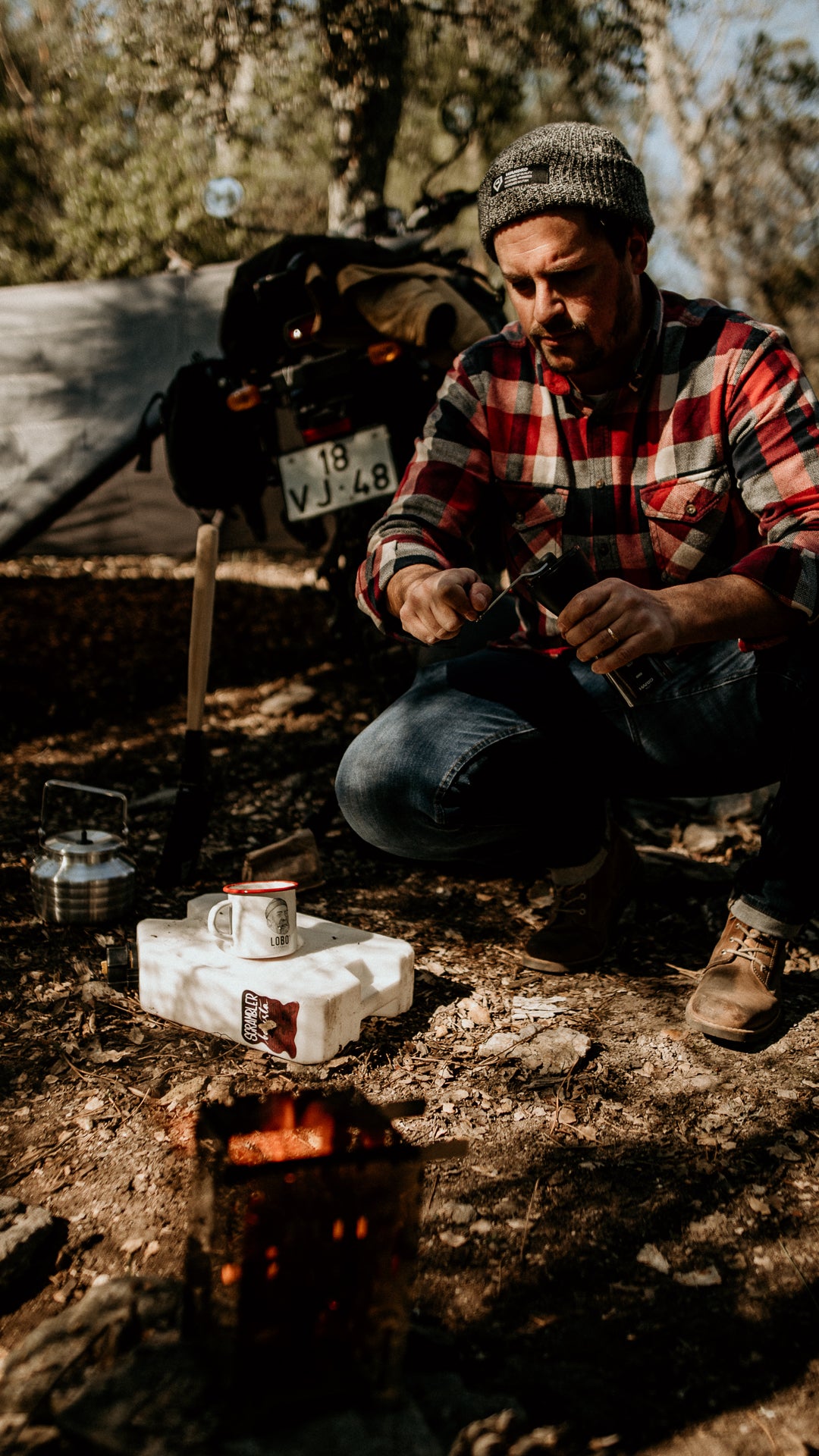 A man wearing a plaid flannel shirt and Lobo Raw Boots preparing coffee outdoors, sitting next to a Lobo Enamel Mug in a forest camping setting.