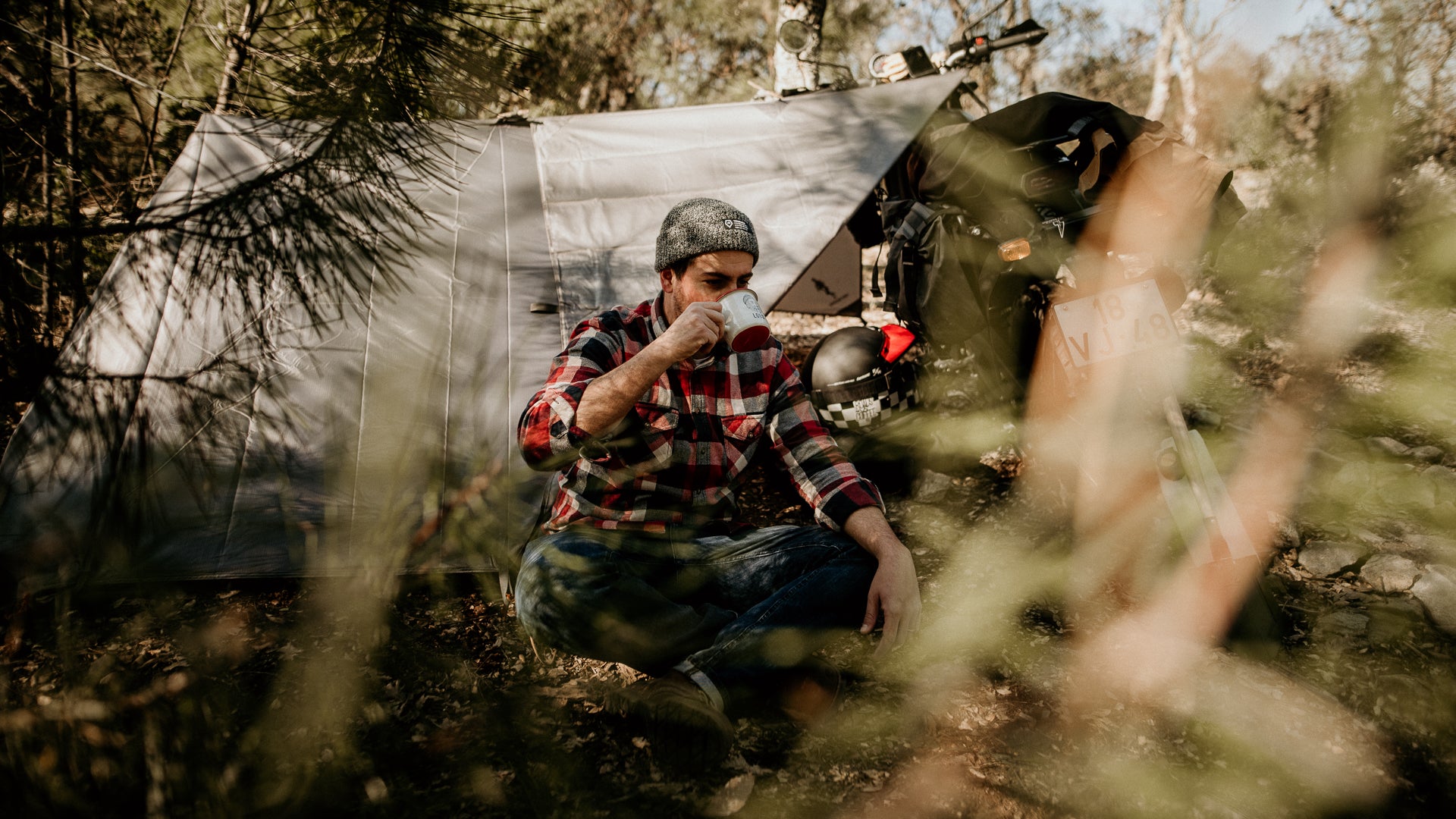A man sitting in front of a grey tarp tent, drinking coffee from a Lobo Enamel Mug, with an adventure motorcycle parked nearby in a forest setting.