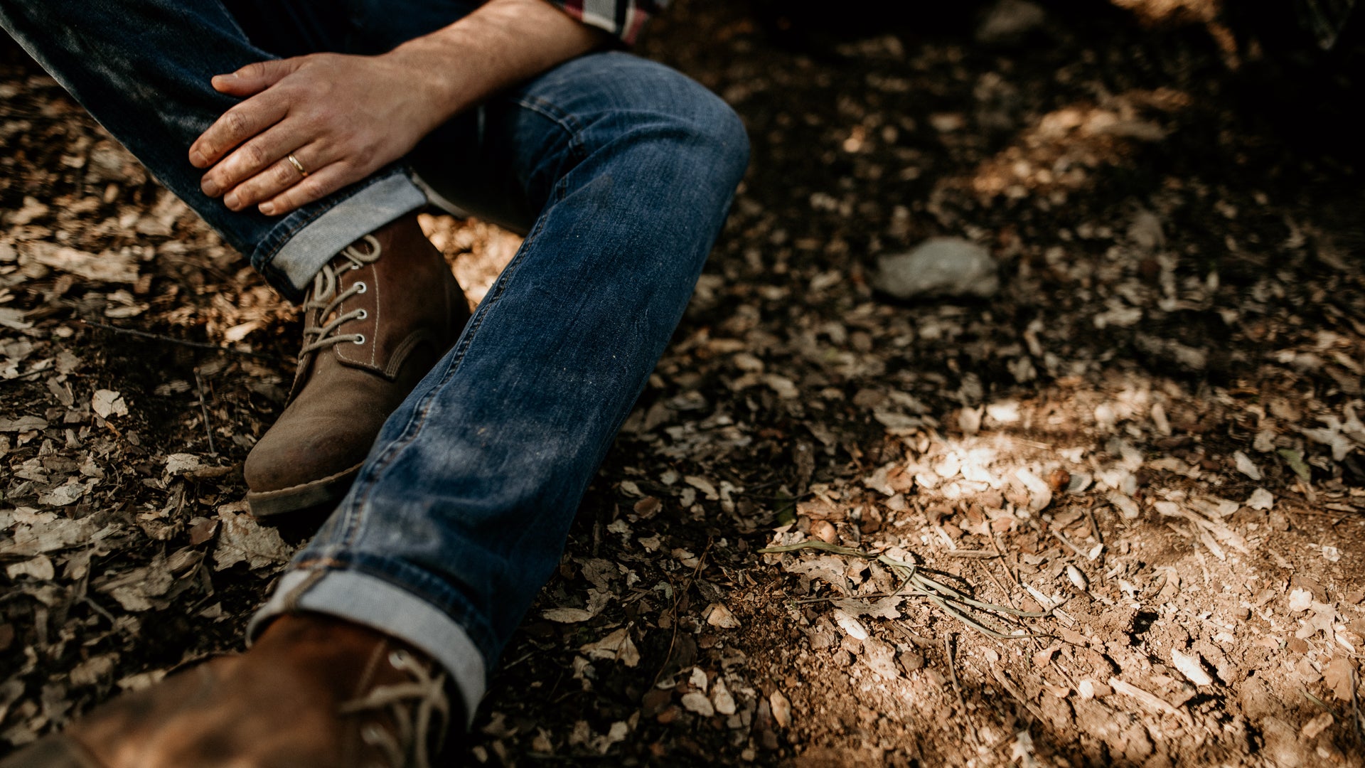 Close-up detail of the Lobo Raw Boots in brown leather, worn with denim jeans on forest ground covered in dry leaves.