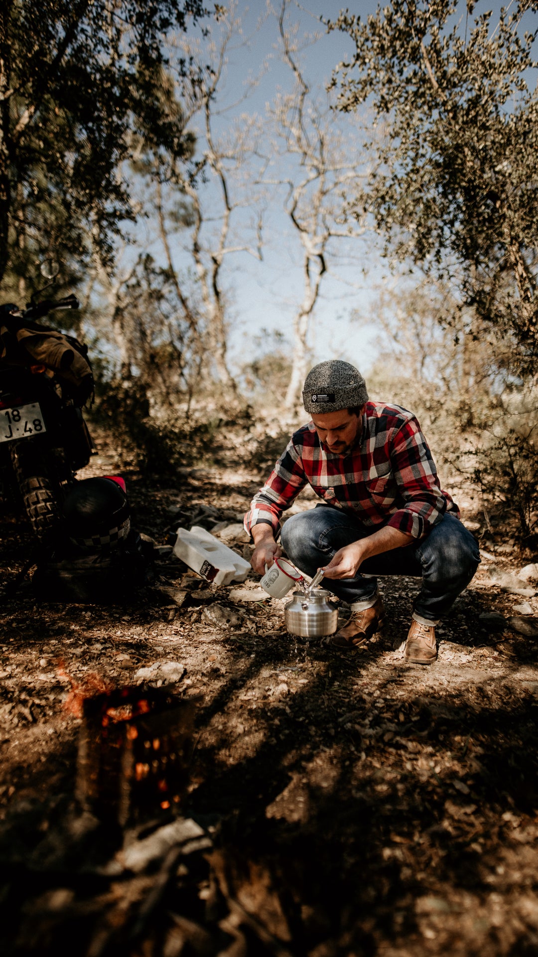 A man wearing Lobo Raw Boots and a beanie, crouching by a campfire pouring water into a kettle, with a Lobo Enamel Mug and an adventure motorcycle in the background of a forest setting.