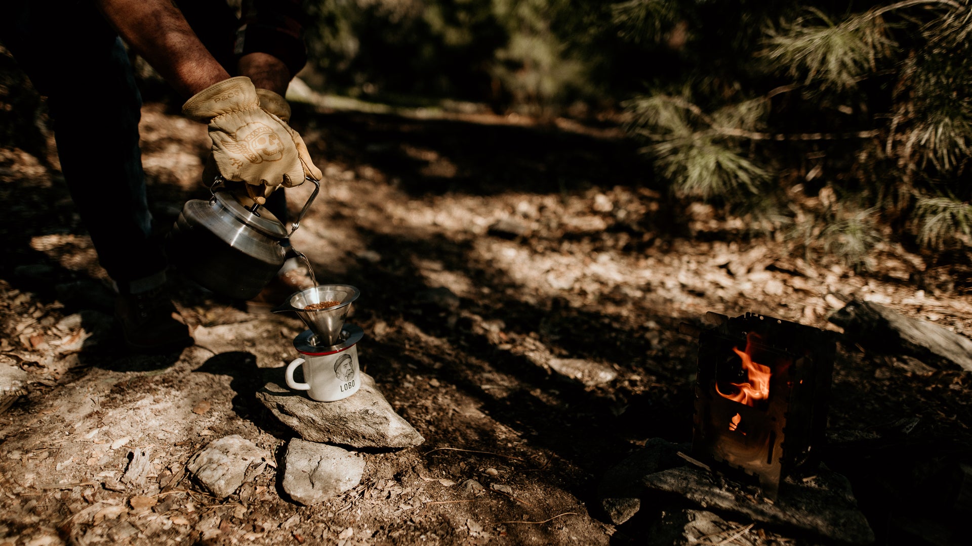 Close-up of coffee being brewed into a Lobo Enamel Mug placed on rocks, with a lit camp stove nearby and leather work gloves in use.