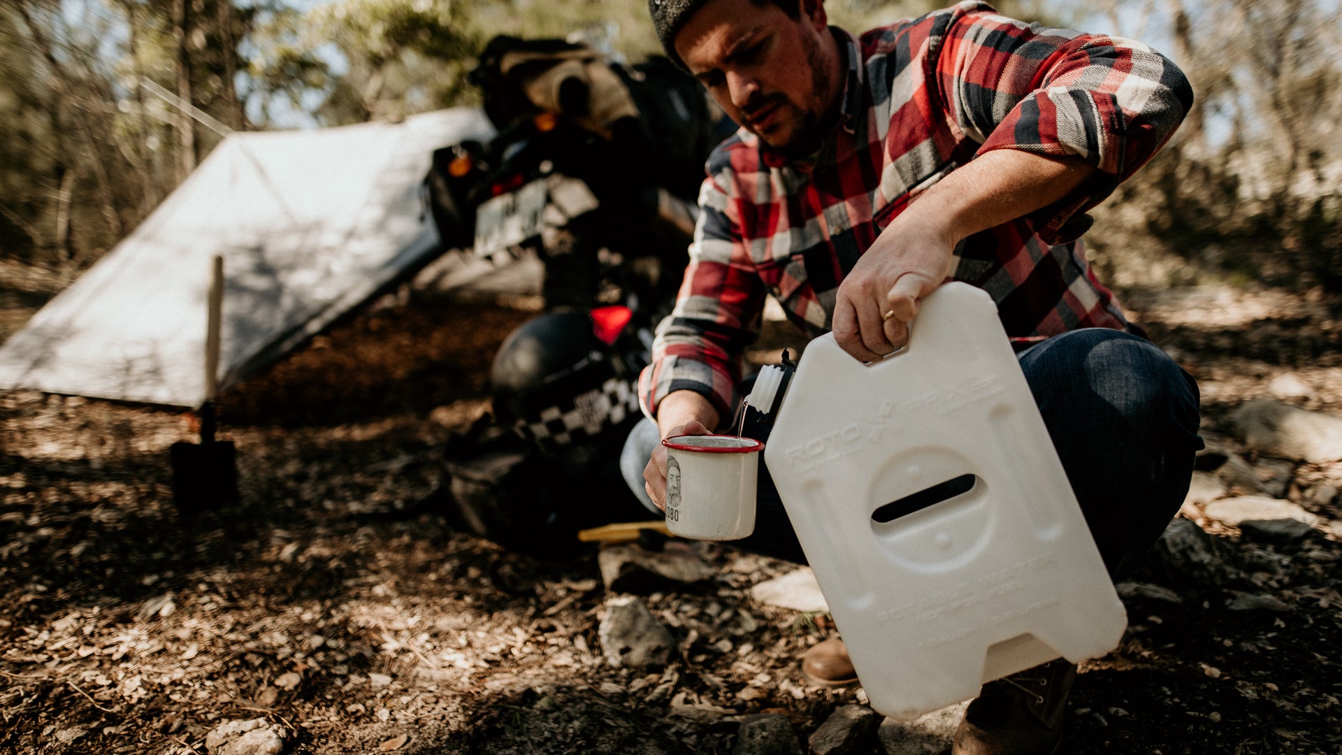A man in a flannel shirt pouring water into a Lobo Enamel Mug, sitting in a campsite with a tent and adventure motorcycle visible.