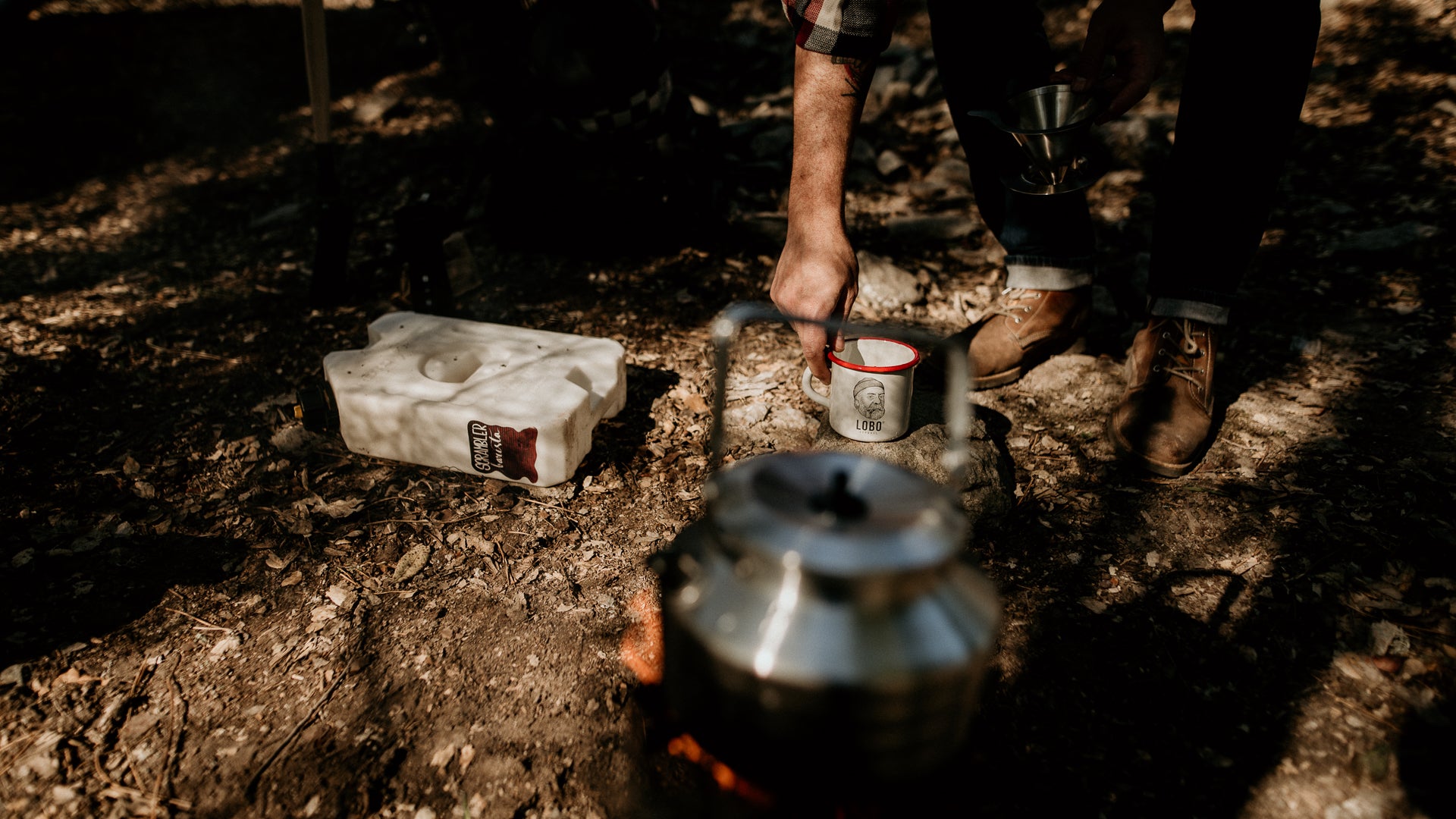 Close-up of leather Lobo Raw Boots and a Lobo Enamel Mug on the forest floor, with a kettle over a campfire in the foreground.