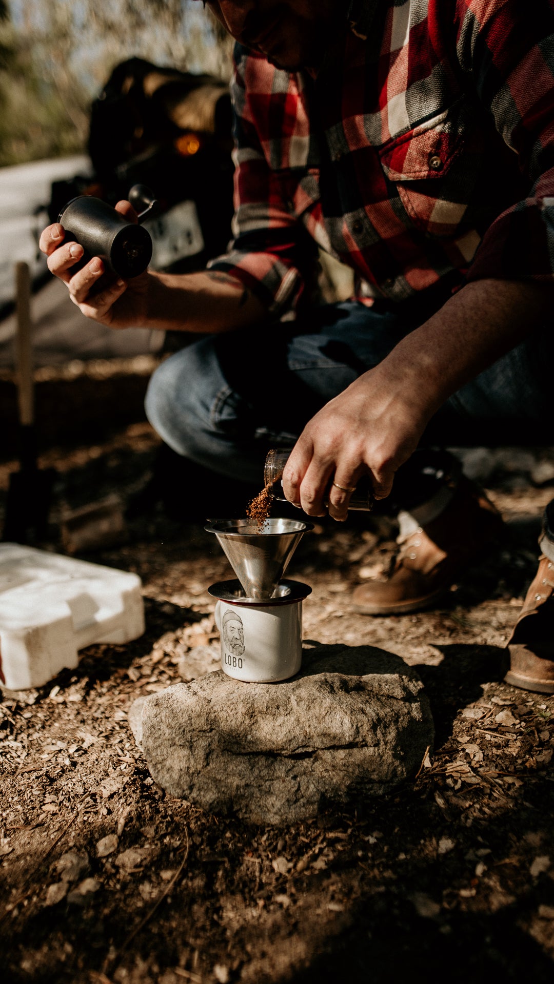 Hands pouring ground coffee into a filter over a Lobo Enamel Mug placed on a rock, with Lobo Raw Boots visible in the background.