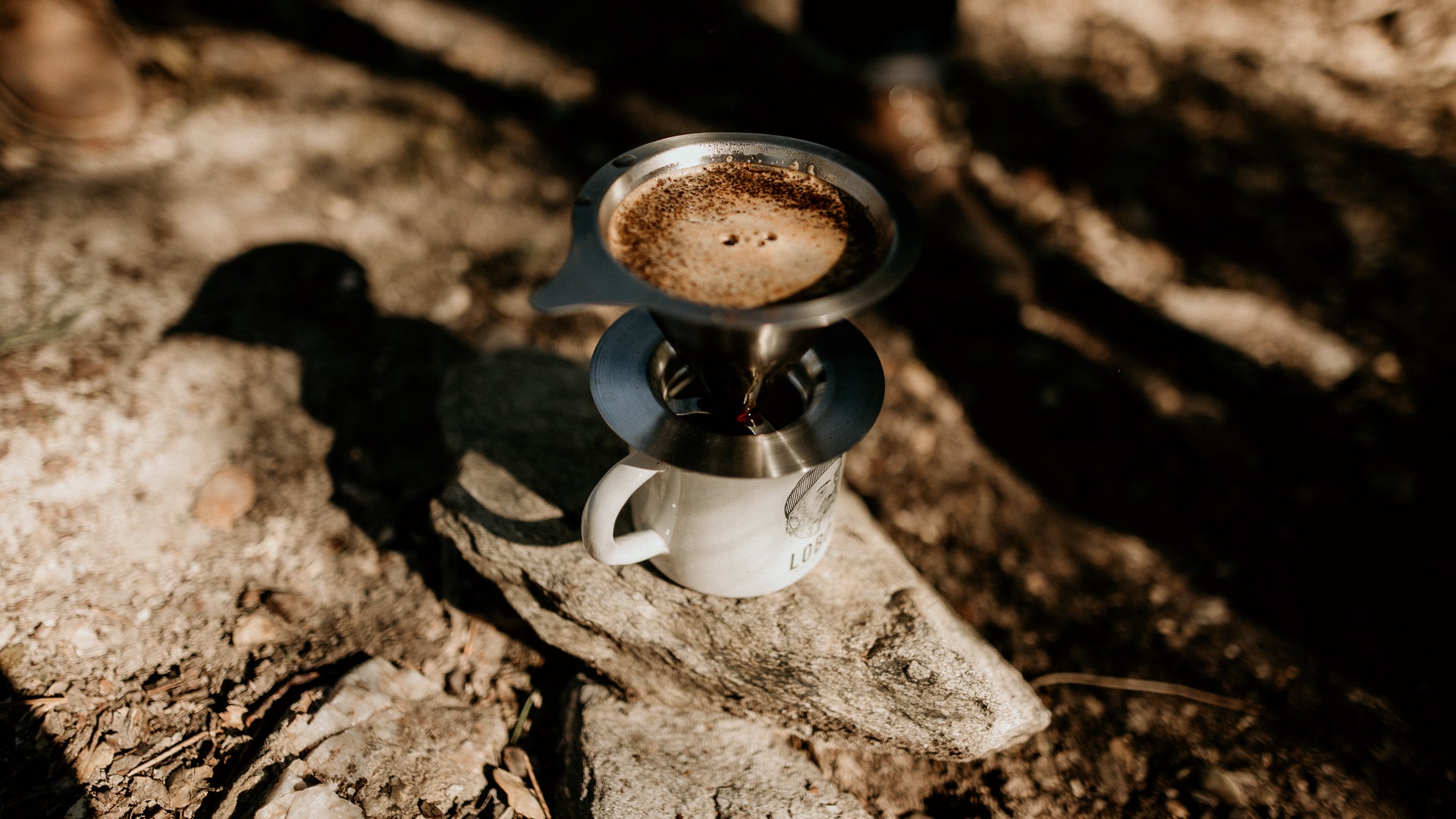 Close-up of a white Lobo Enamel Mug resting on a rock, with a metal coffee dripper on top brewing coffee in an outdoor setting.