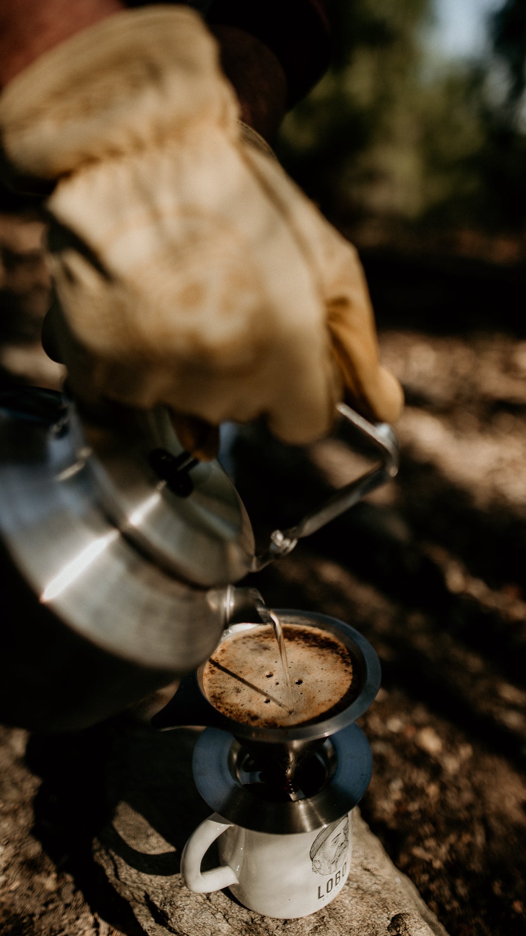 Close-up detail of hot water being poured from a kettle into coffee, during an outdoor brewing ritual with leather gloves.