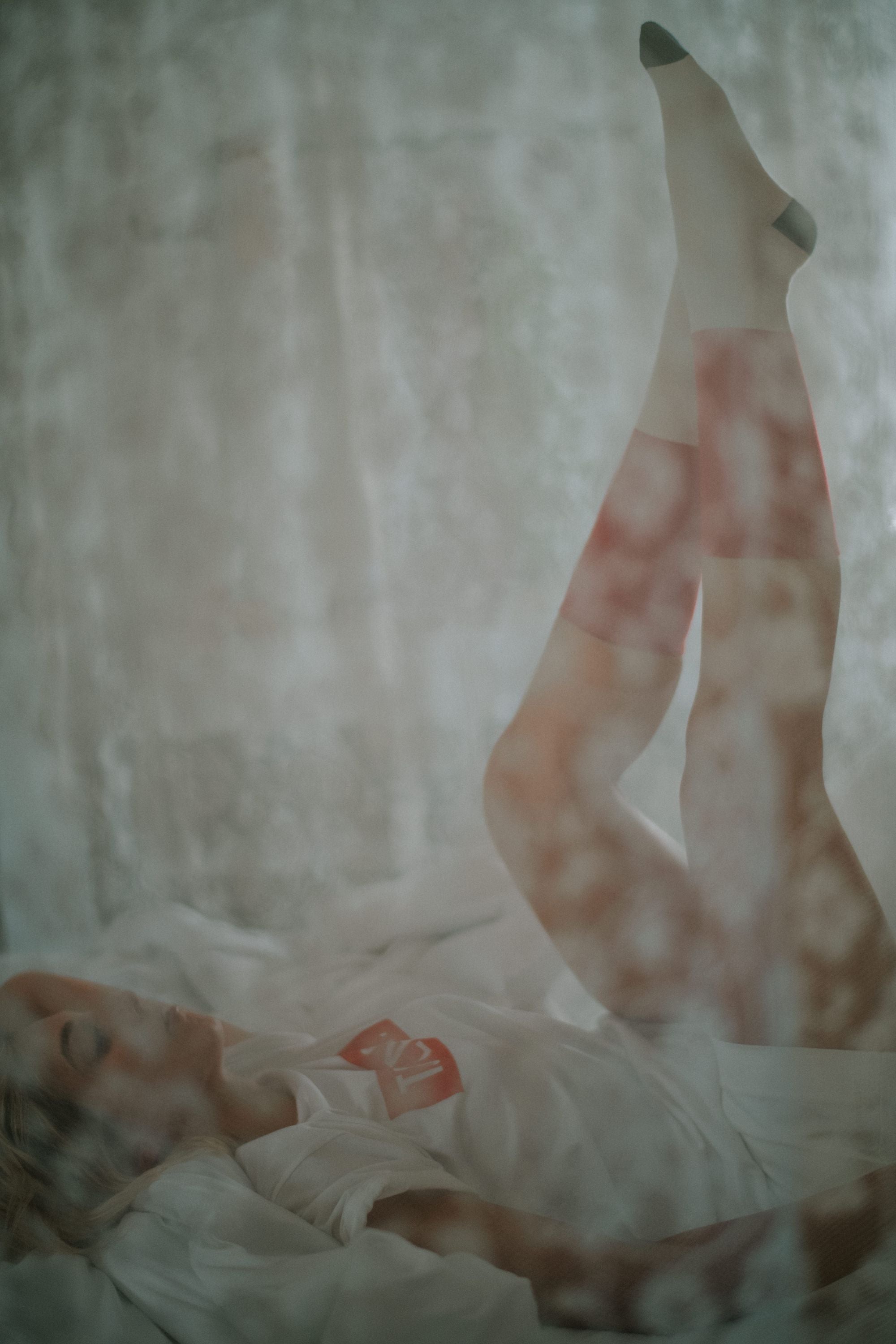 Woman lying on a white bed with legs raised, seen through a soft light effect, wearing Lobo Classic Red Socks.
