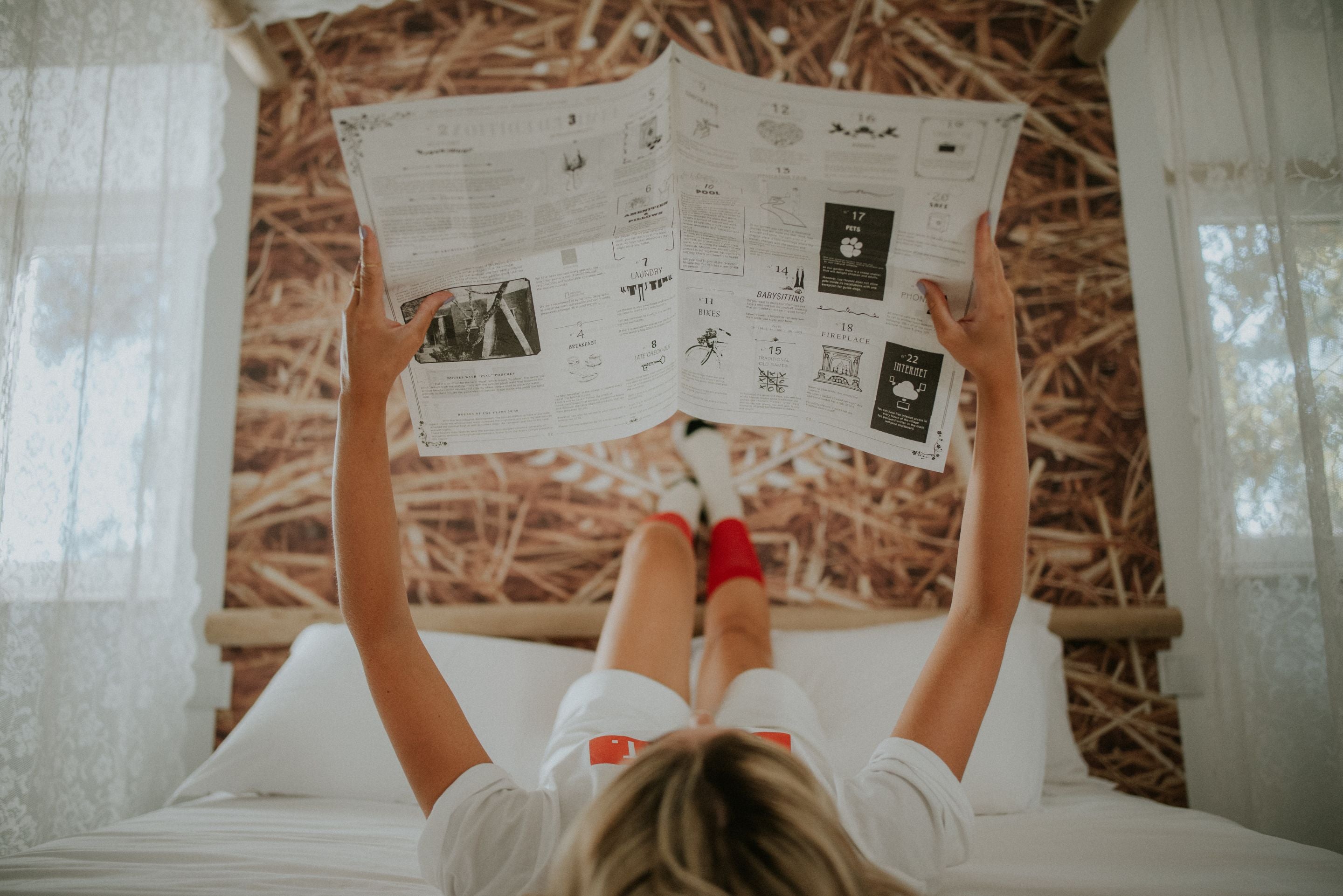 Overhead view of a woman lying in bed wearing the white Lobo Too Last t-shirt and Lobo Classic Red Socks while reading an illustrated newspaper.