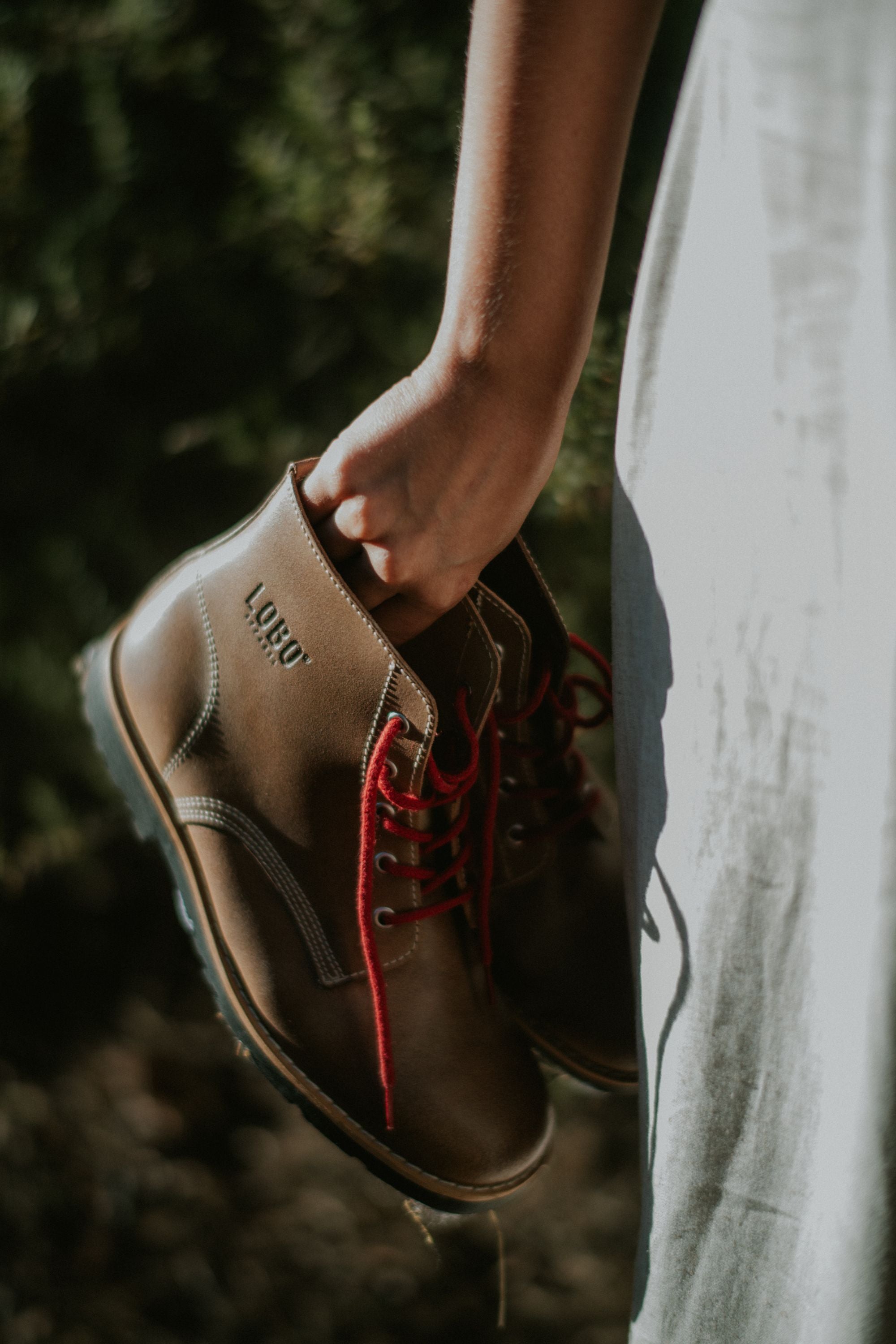 A hand holding a pair of brown leather Lobo Raw Boots with red laces, highlighting the engraved LOBO logo on the leather.