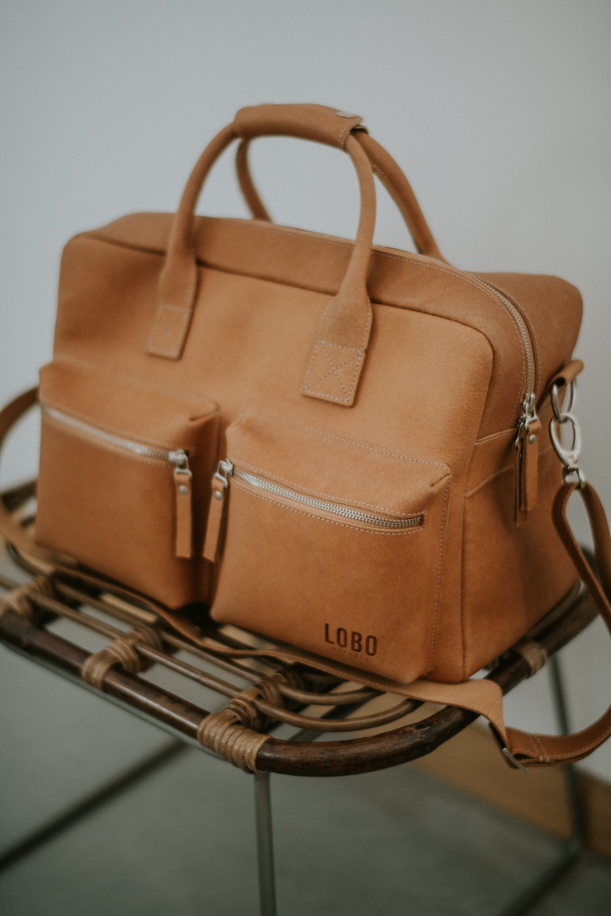 Brown leather travel bag Lobo Leather Suitcase featuring front pockets and metal zippers, resting on a wicker bench.