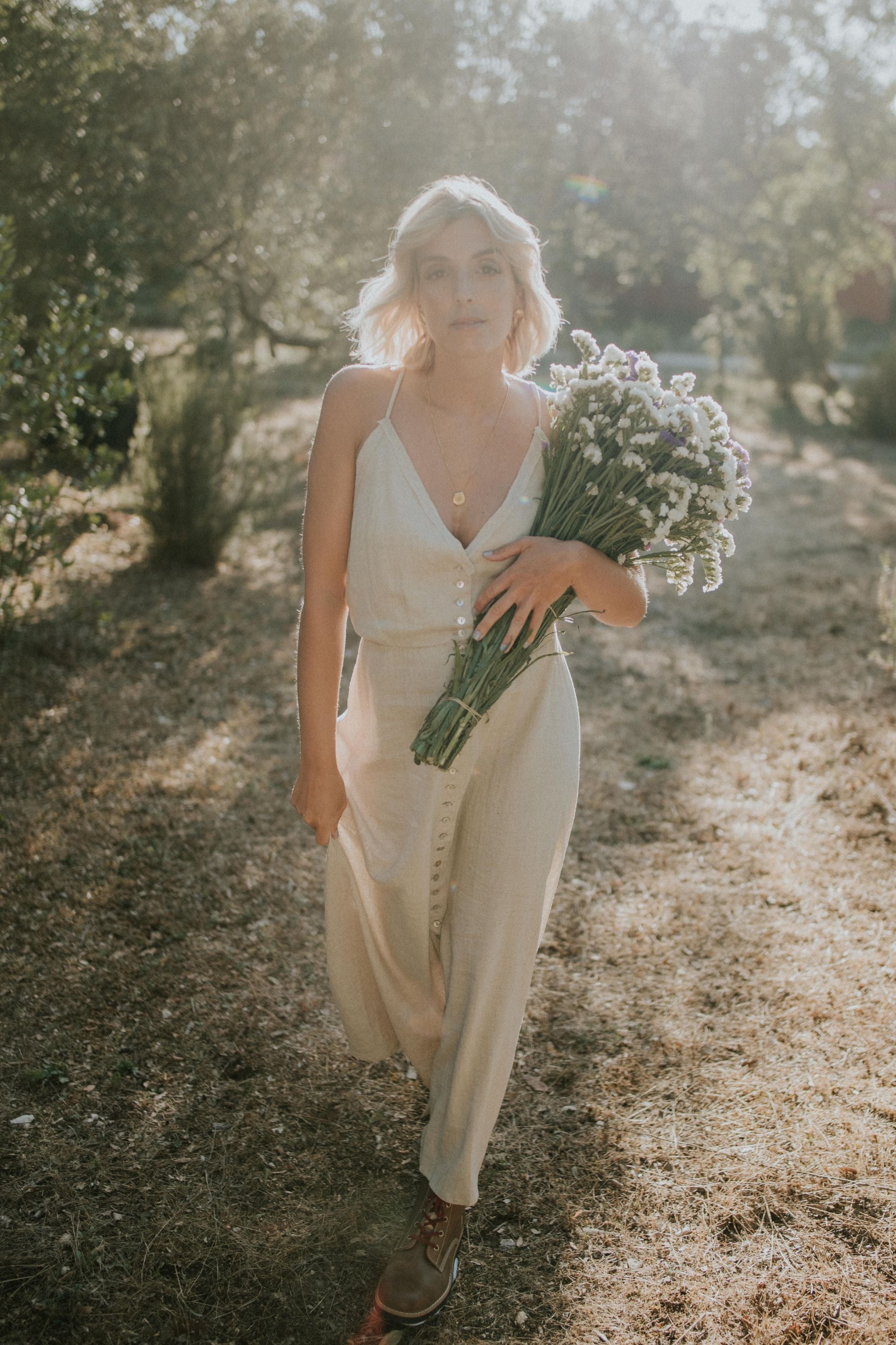 Blonde model walking forward in a field, wearing a linen dress and Lobo Raw Boots, holding a bouquet of wildflowers.