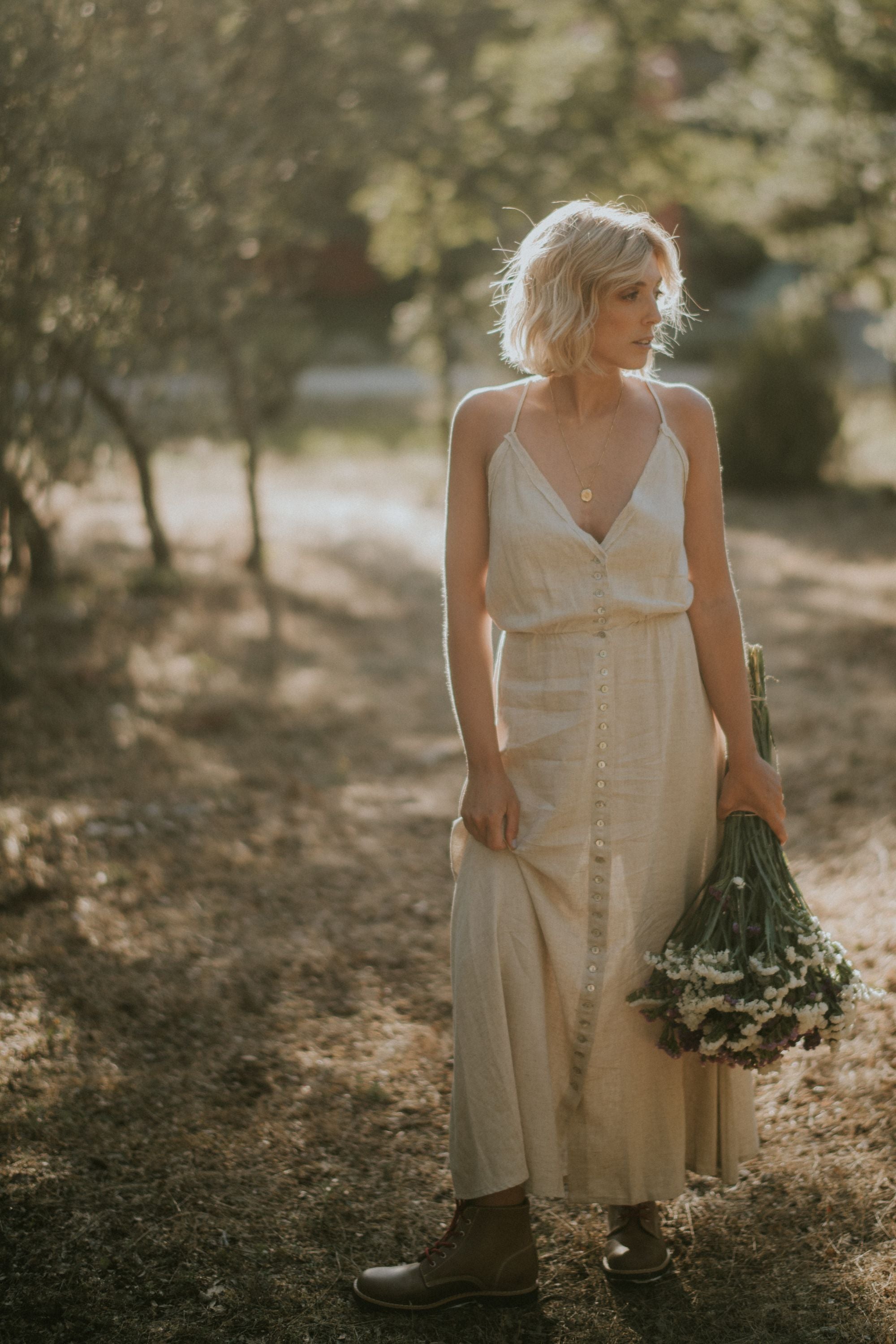 Blonde woman in a sunlit field, wearing a beige dress and Lobo Raw Boots leather boots while holding a bouquet of flowers.