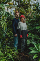 A couple in a lush tropical garden; the man wears the Manuel Brown Beanie wool hat and a blue knit sweater, while the woman wears a red beanie and two-tone sweater.