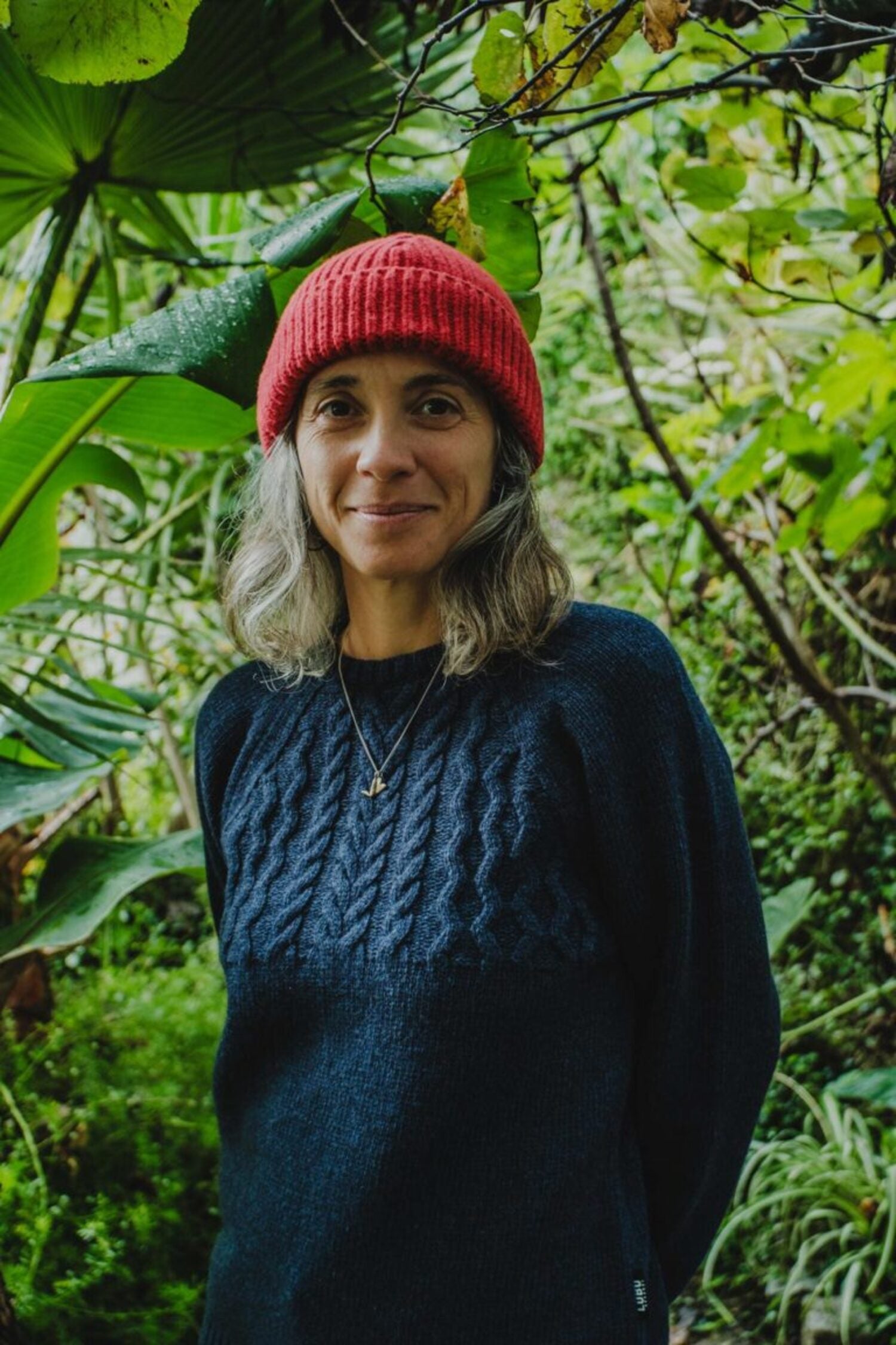 Woman in a lush garden wearing the red Manuel Red Beanie and a dark blue knit sweater.