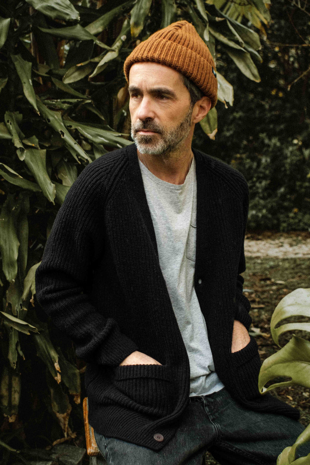Founder Bruno Pereira posing in a natural outdoor setting wearing the brand's products: the Lobo Black Wool Cardigan, the Lobo Grey T-shirt with a pocket, and the Manuel Brown Beanie.