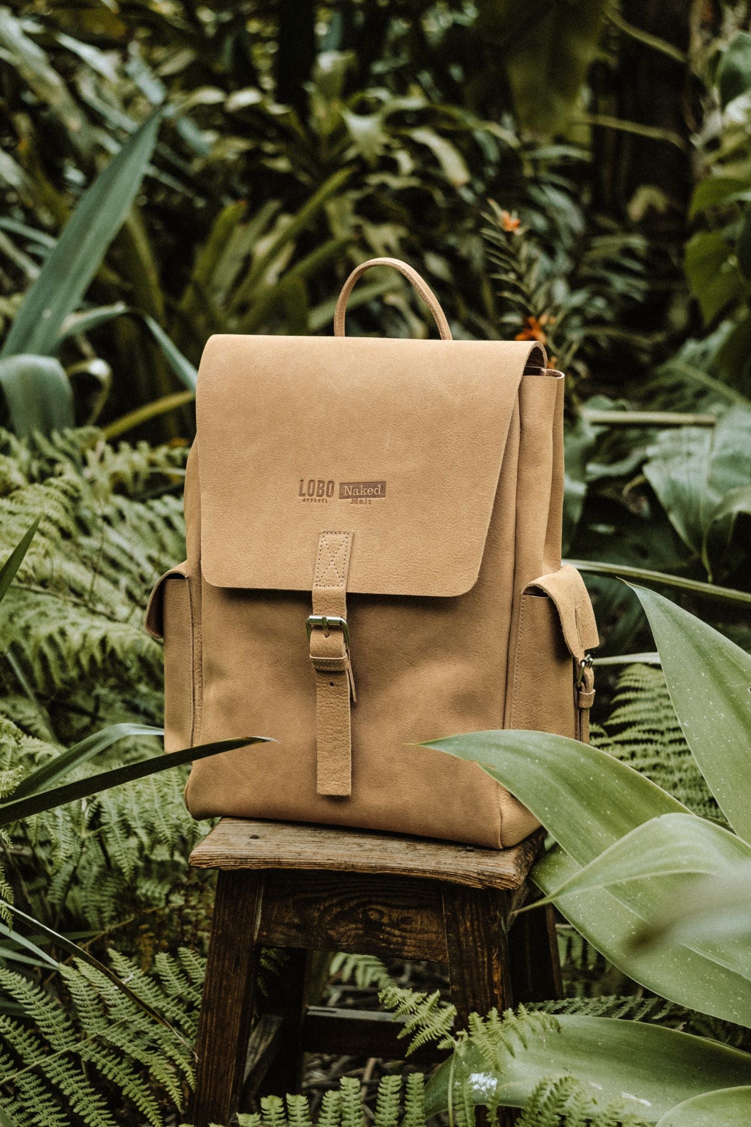Lobo x Naked Malt brown leather backpack placed on a wooden stool, surrounded by tropical green vegetation and ferns. Highlighting the side pockets and embossed logos.