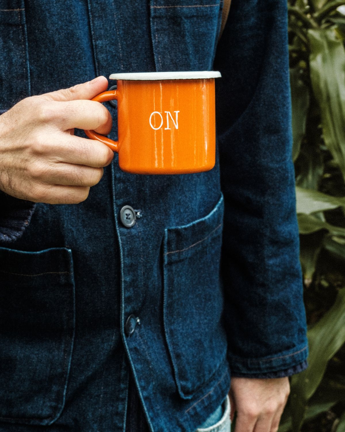 Close-up of a man in a denim jacket holding the orange Lobo Apparel x Naked Malt collab enamel mug.