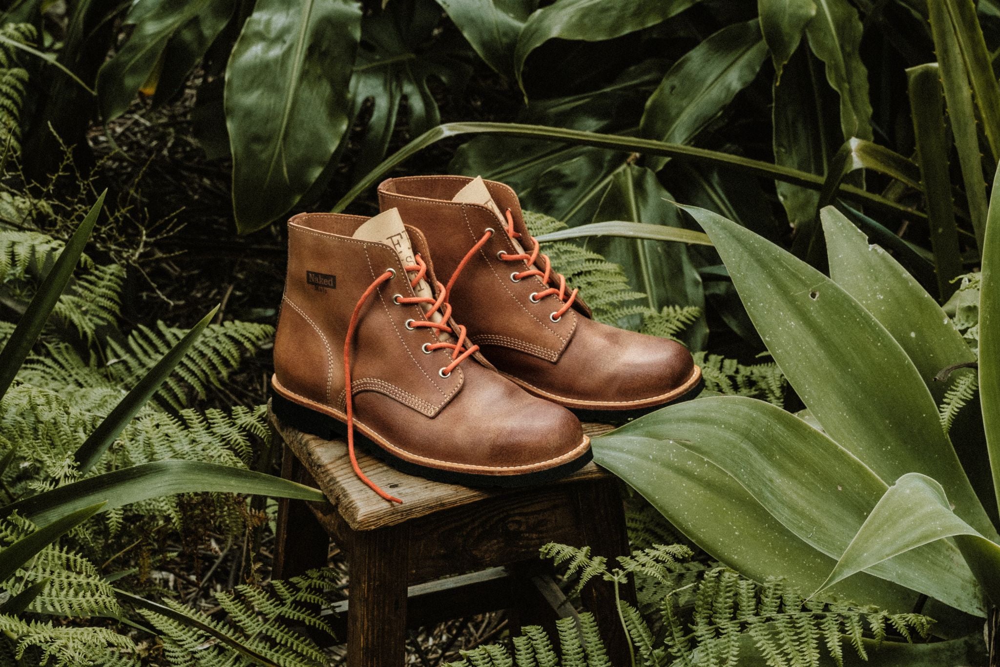 A pair of handmade Lobo Apparel x Naked Malt boots displayed outdoors amidst lush green ferns and tropical plants.