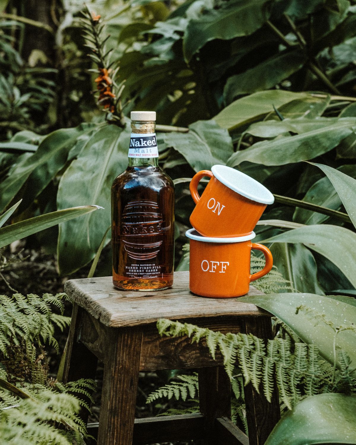 A bottle of Naked Malt whisky next to two orange enamel mugs from the Lobo x Naked Malt Collab Enamel Mug collection, stacked with "ON" and "OFF" inscriptions, in a natural setting.