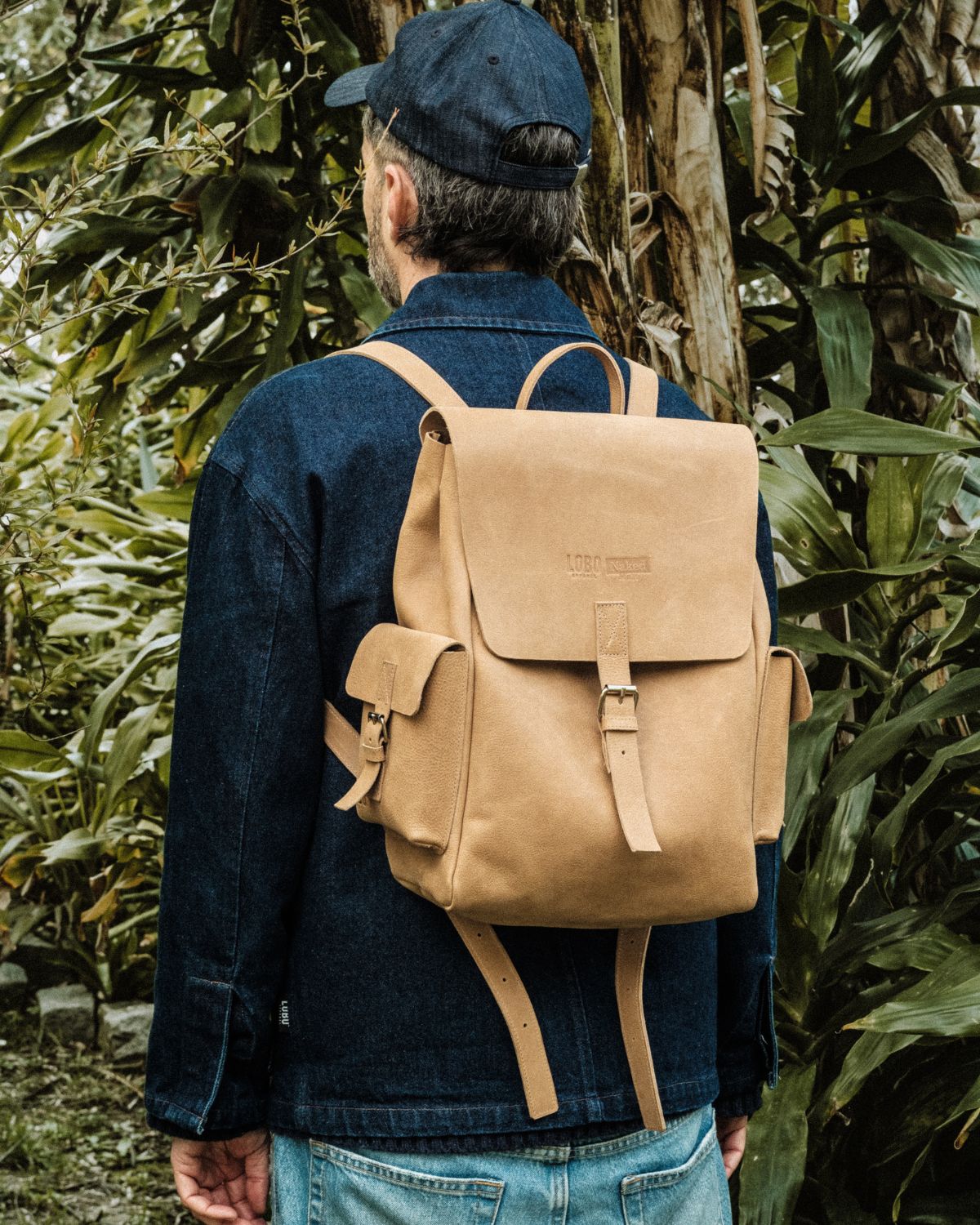 Rear view of a man wearing the Lobo x Naked Malt Collab Backpack, a sand-colored leather backpack with side pockets, featured in an outdoor look with a blue jacket.