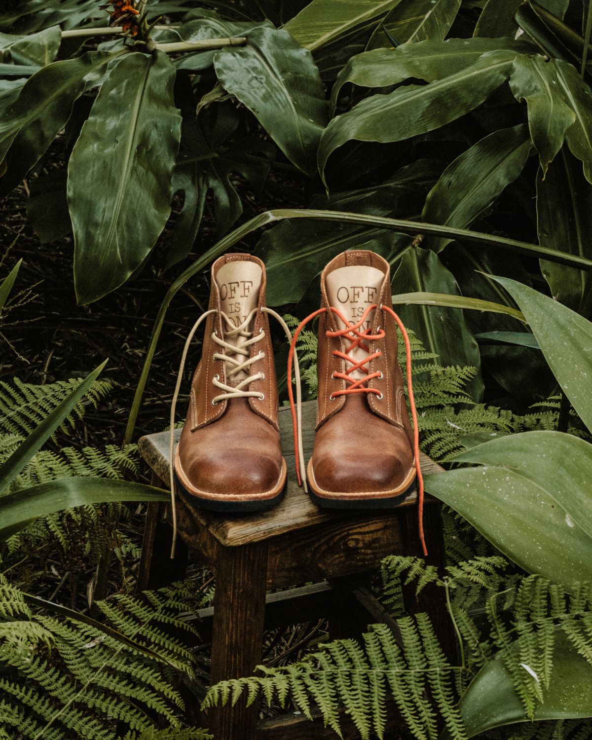 A pair of brown leather Lobo x Naked Malt Collab Raw Boots placed on a wooden stool in a tropical forest setting, featuring beige and orange laces with the "OFF IS ON" inscription.