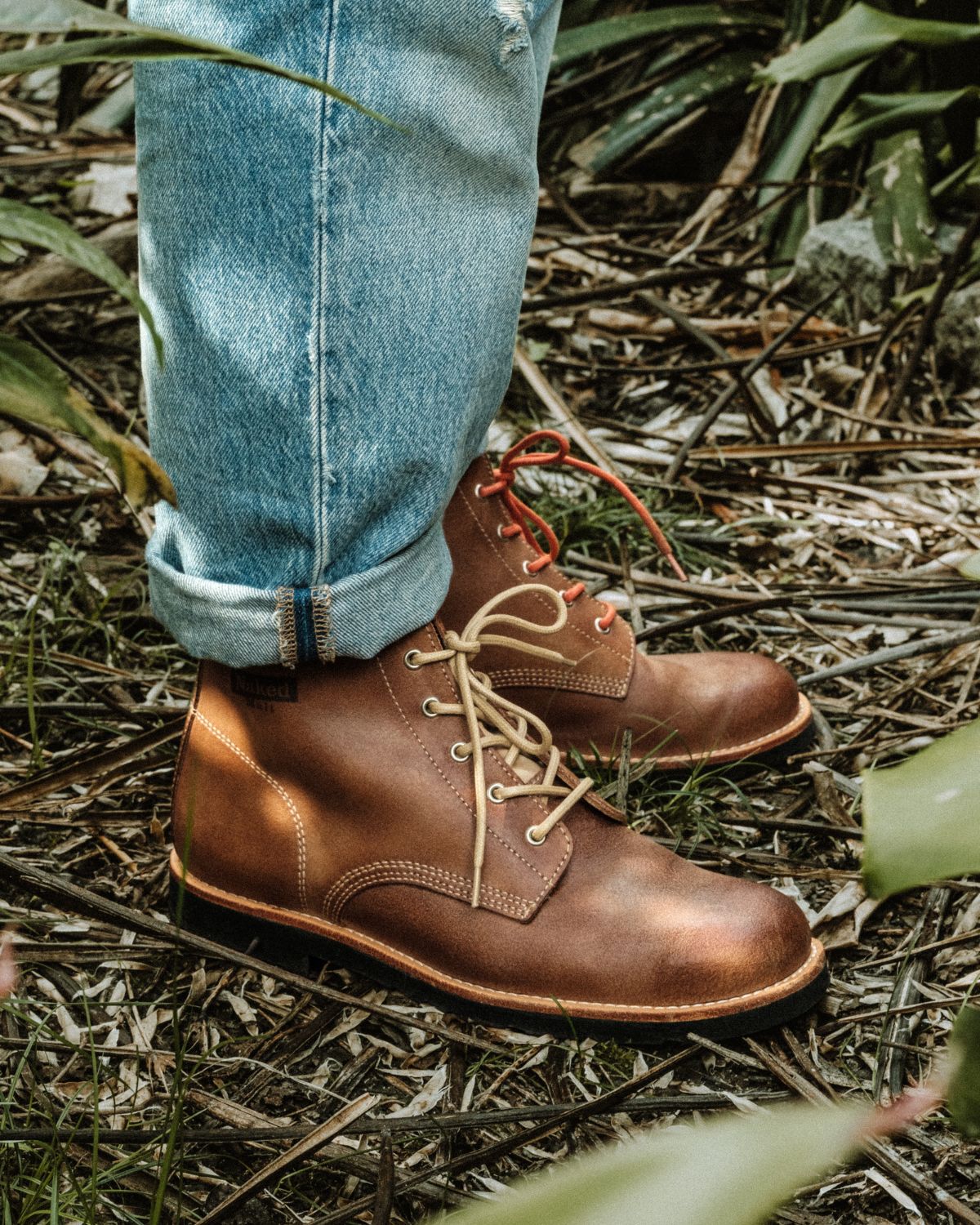 Lobo x Naked Malt Collab Raw Boots in brown leather with beige and orange laces in an outdoor setting.
