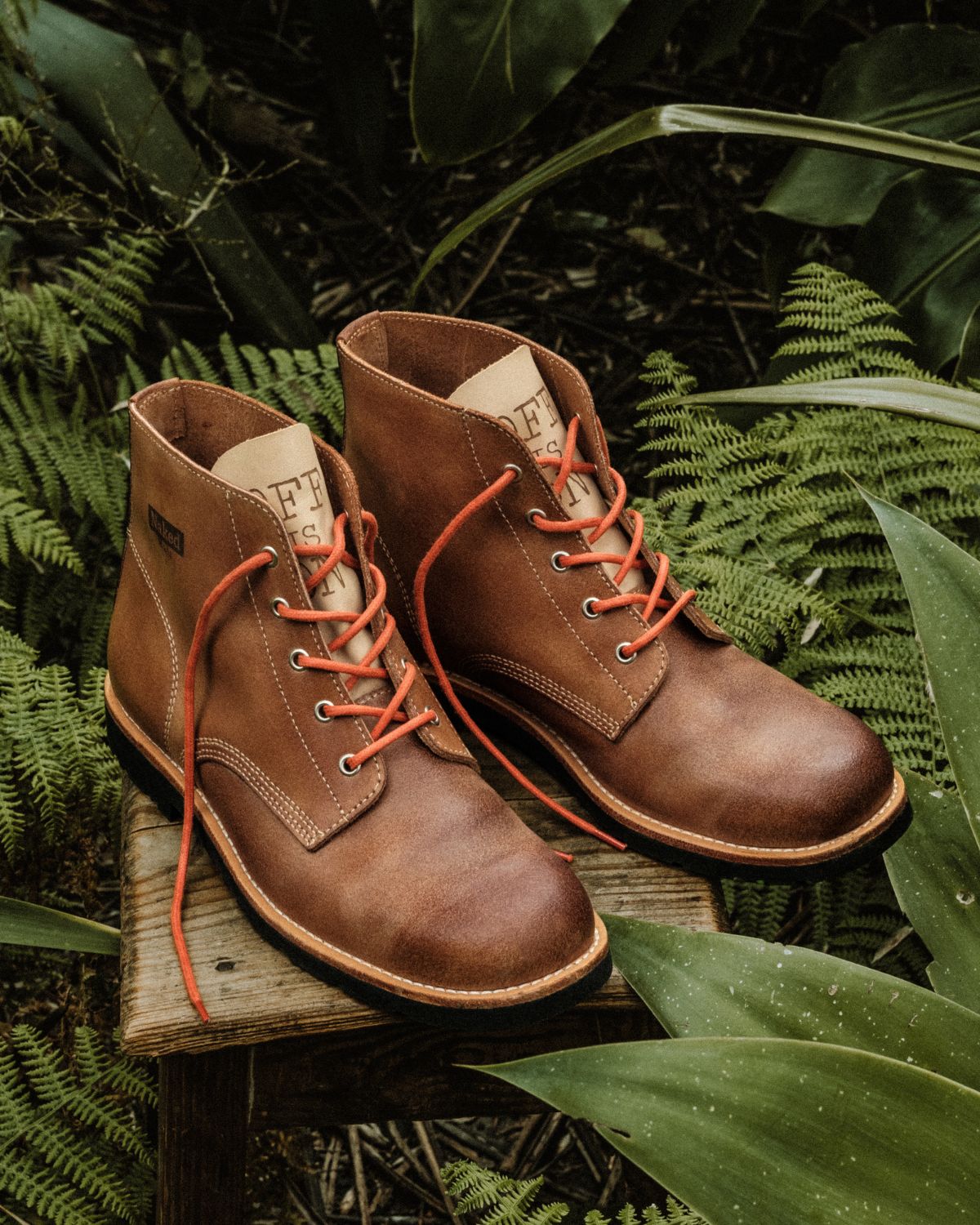 Side view of a pair of brown leather Lobo x Naked Malt Collab Raw Boots with orange laces, set on a wooden stool in a natural forest environment.