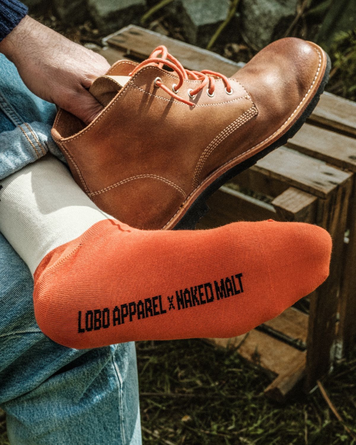 Close-up of the sole of the foot wearing Lobo x Naked Malt Collab Socks, displaying the phrase "LOBO APPAREL X NAKED MALT", with the Lobo x Naked Malt Collab Raw Boots in the background.