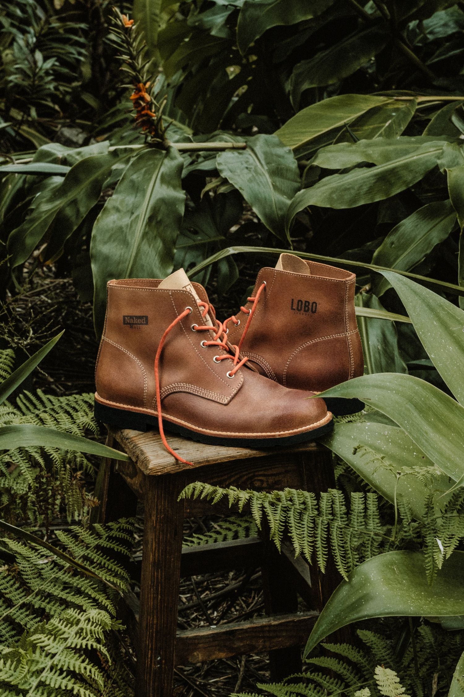 Handcrafted leather boots from the Lobo x Naked Malt collaboration, inspired by traditional Portuguese work boots, set against a lush green foliage background.