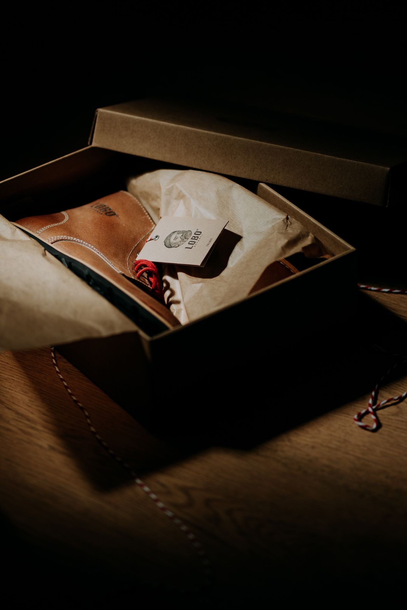 Lobo Raw Boots inside an open cardboard box, with a tag showing the logo of a bearded man with a beanie and a pipe.