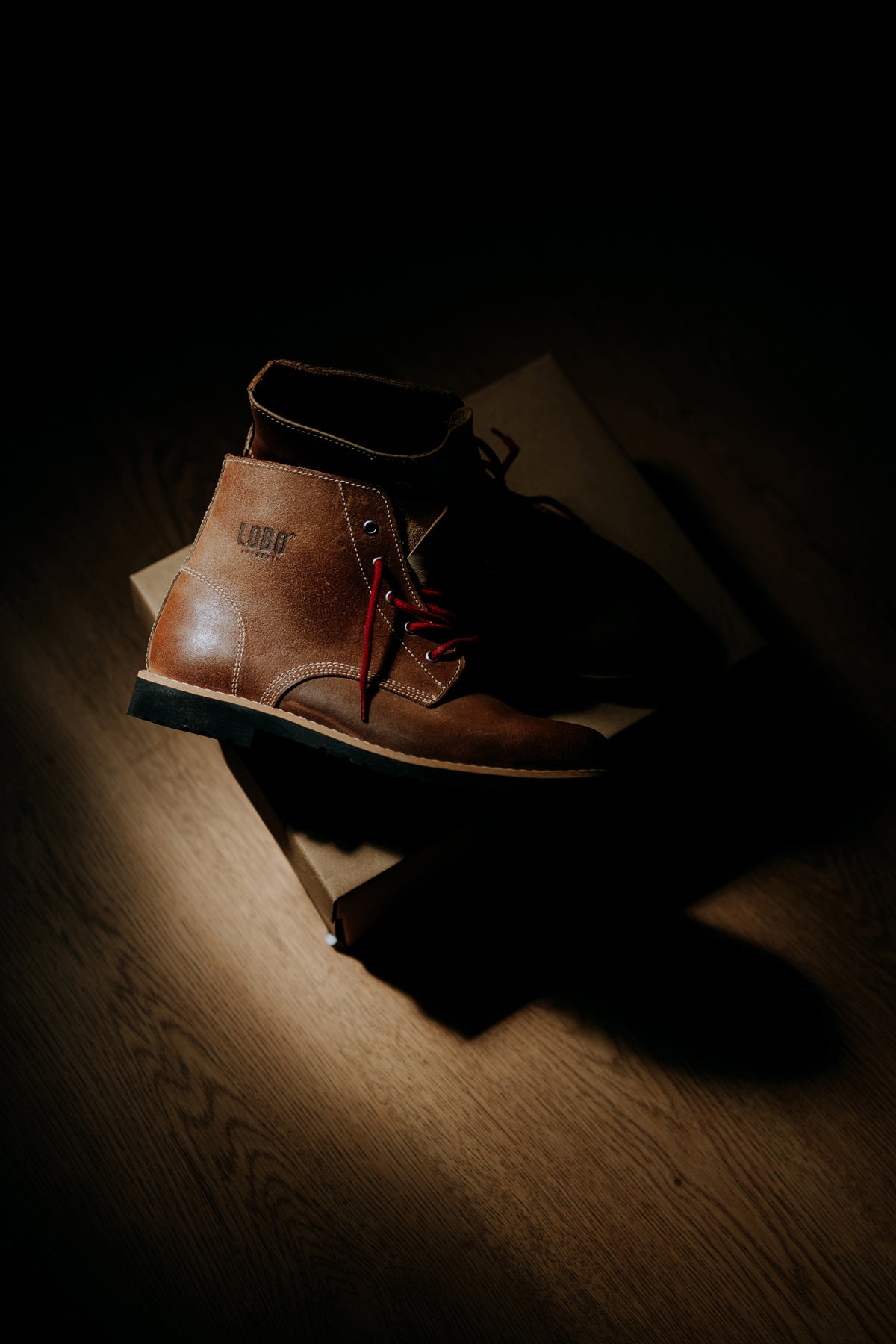 Pair of brown leather Lobo Raw Boots with red laces, side-lit while resting on their original cardboard box.