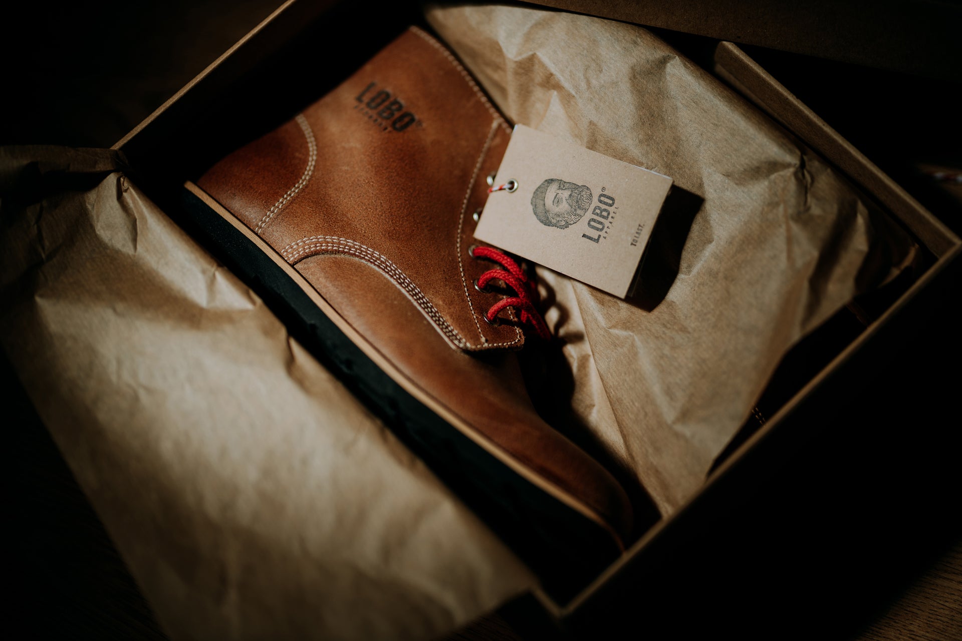 Lobo Raw Boots brown leather boot inside its original box, wrapped in tissue paper, with a hangtag featuring the logo of a bearded man with a beanie and a pipe.