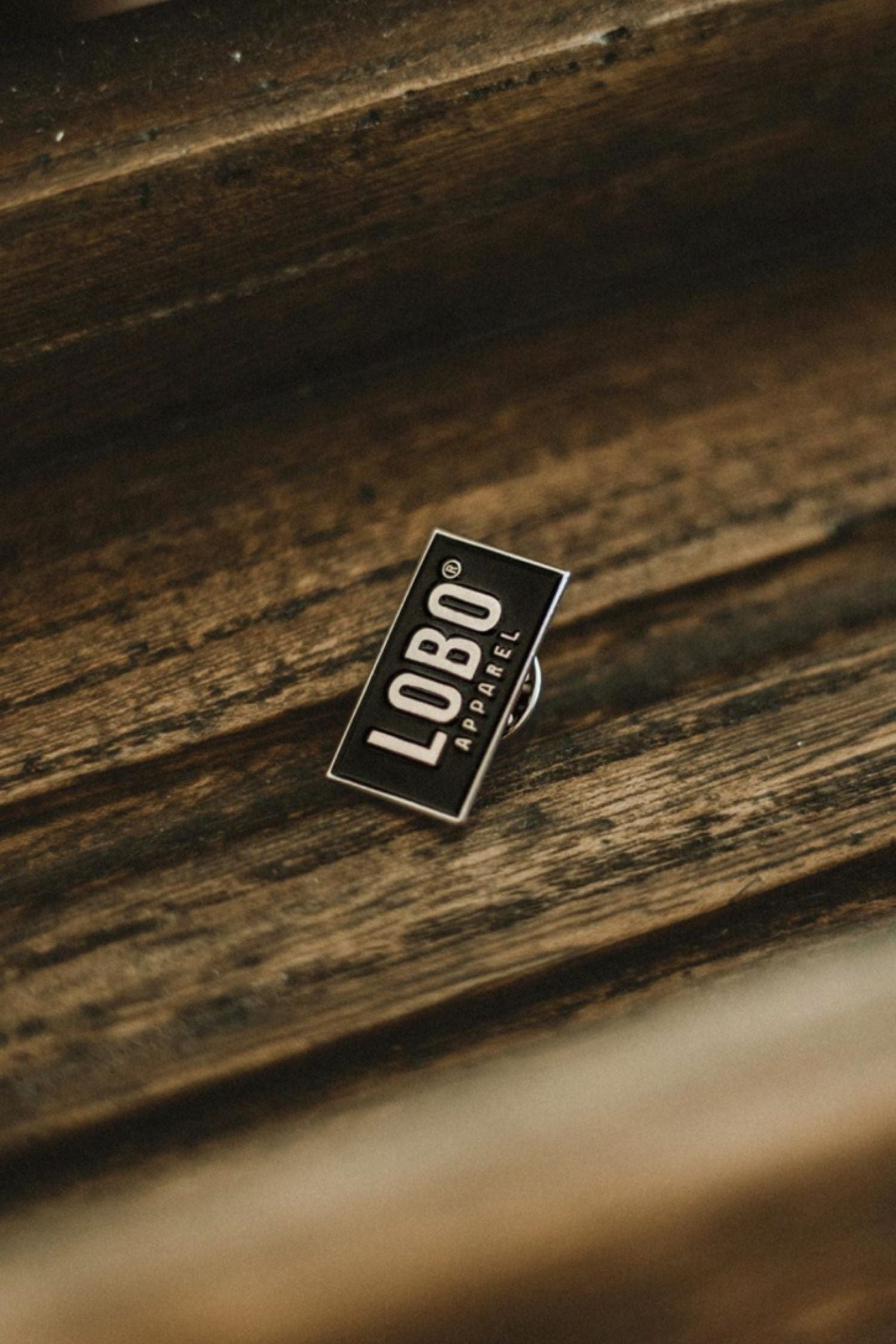 Rectangular black Lobo Apparel metal pin with white "LOBO APPAREL" text, resting on a wooden surface.