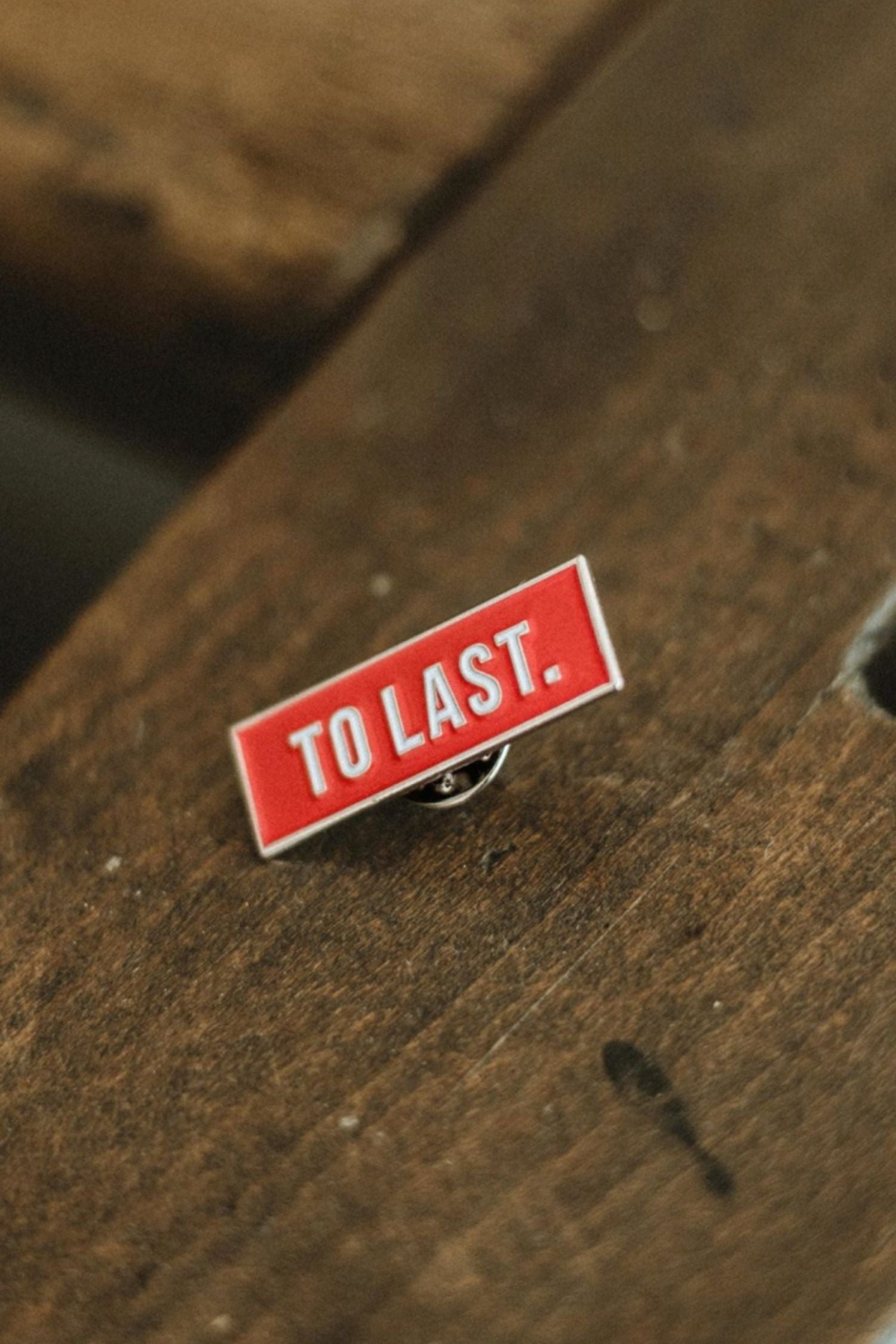 Rectangular Lobo To Last metal pin with red background and white text "TO LAST." resting on wood.