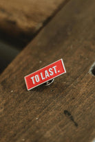 Rectangular Lobo To Last metal pin with red background and white text "TO LAST." resting on wood.