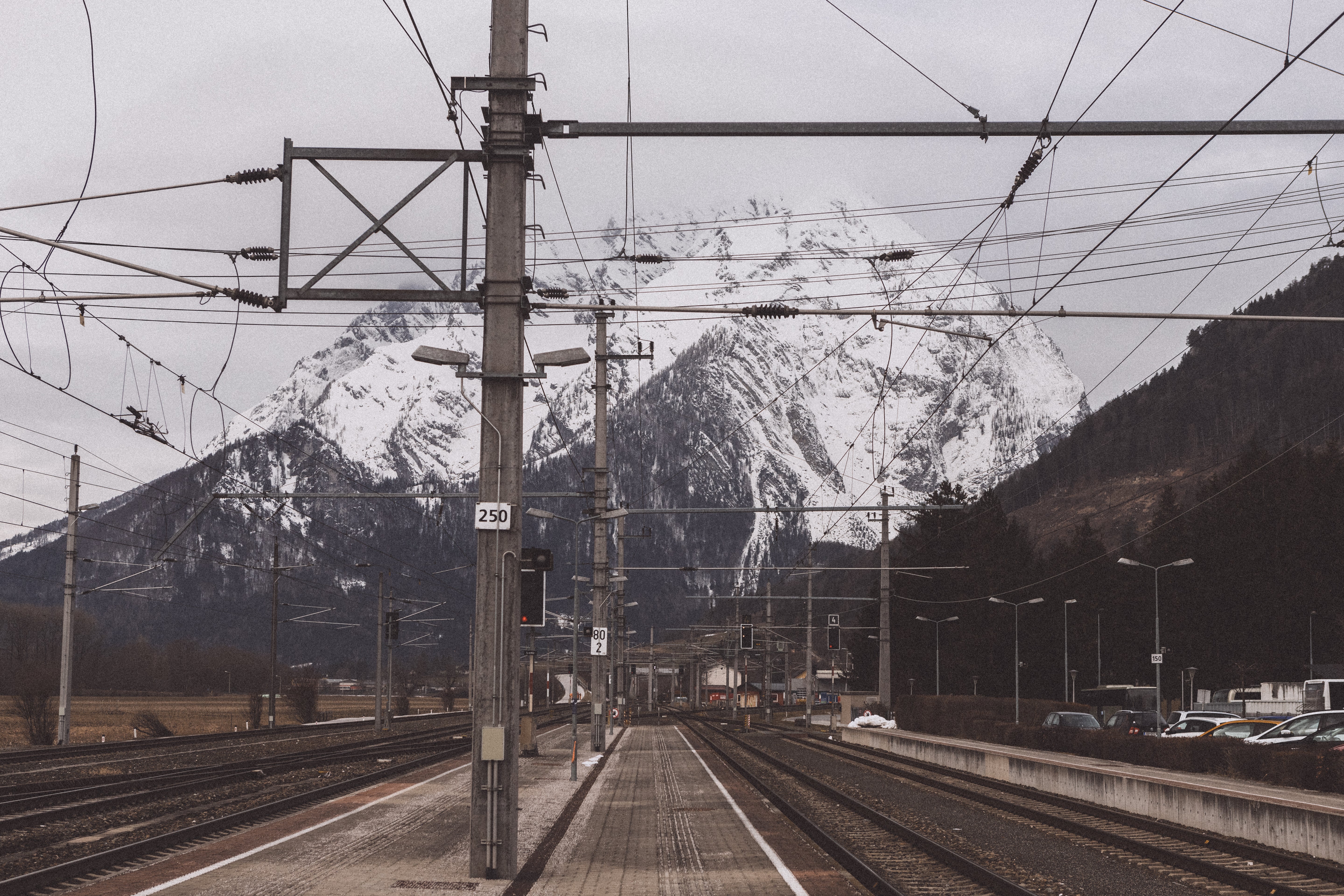 Hallstatt train station with tracks and overhead lines in the foreground, framing a massive snowy mountain under a cloudy sky.