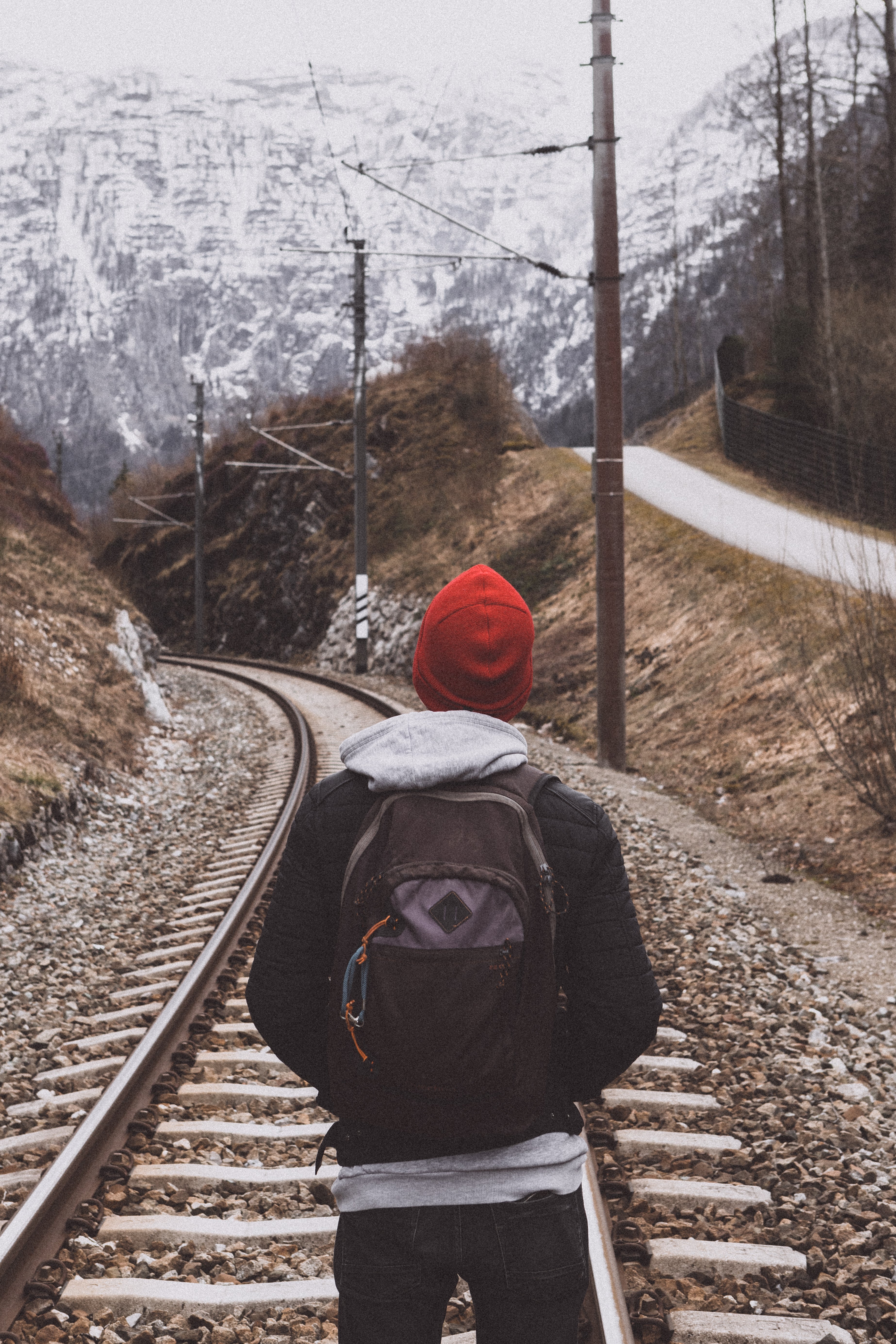 Traveler with a backpack and red beanie walks along a winding railway line through the Austrian mountains.