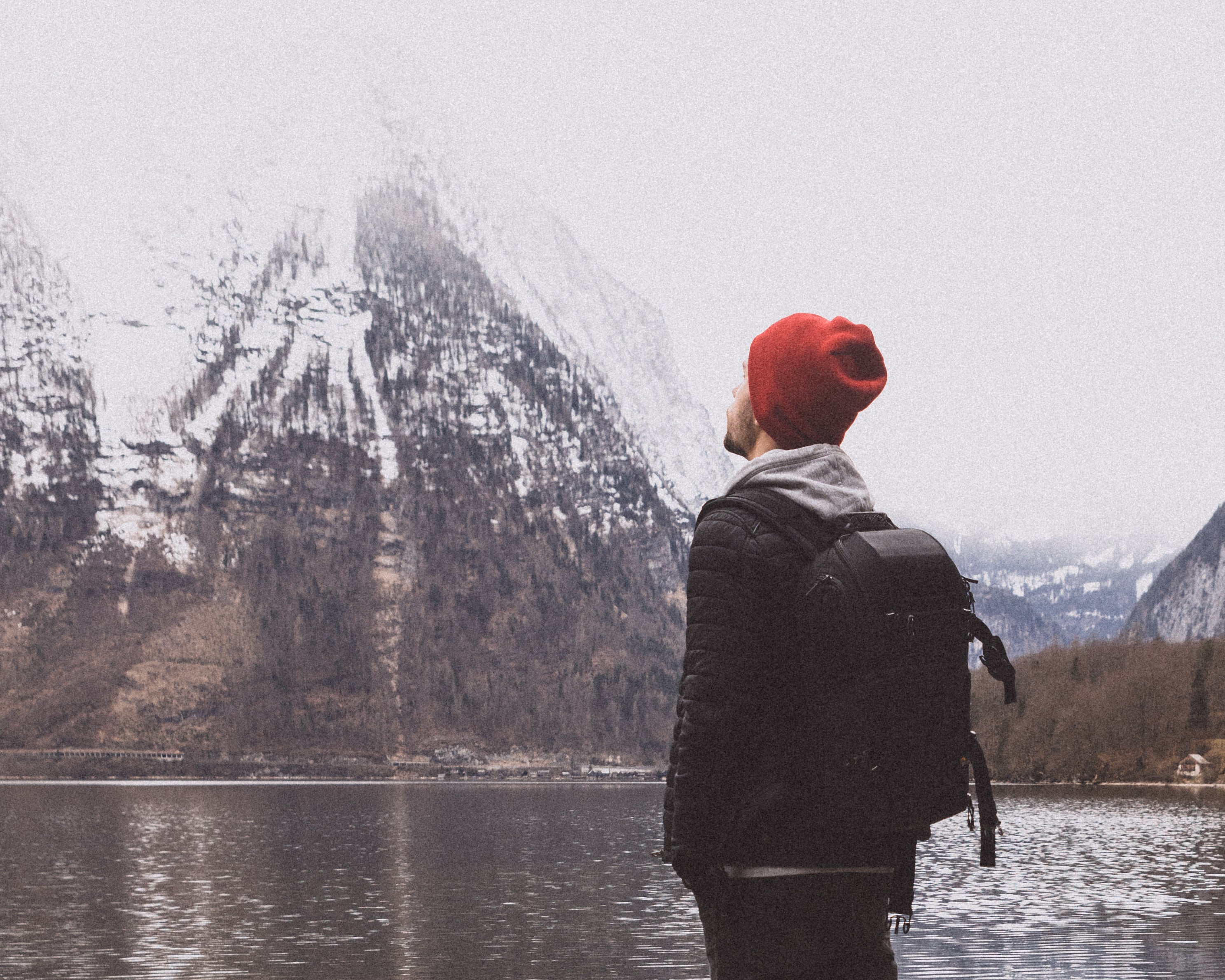 Profile portrait of an explorer wearing a red beanie and technical backpack, looking up at a towering, snowy mountain peak by the lake.