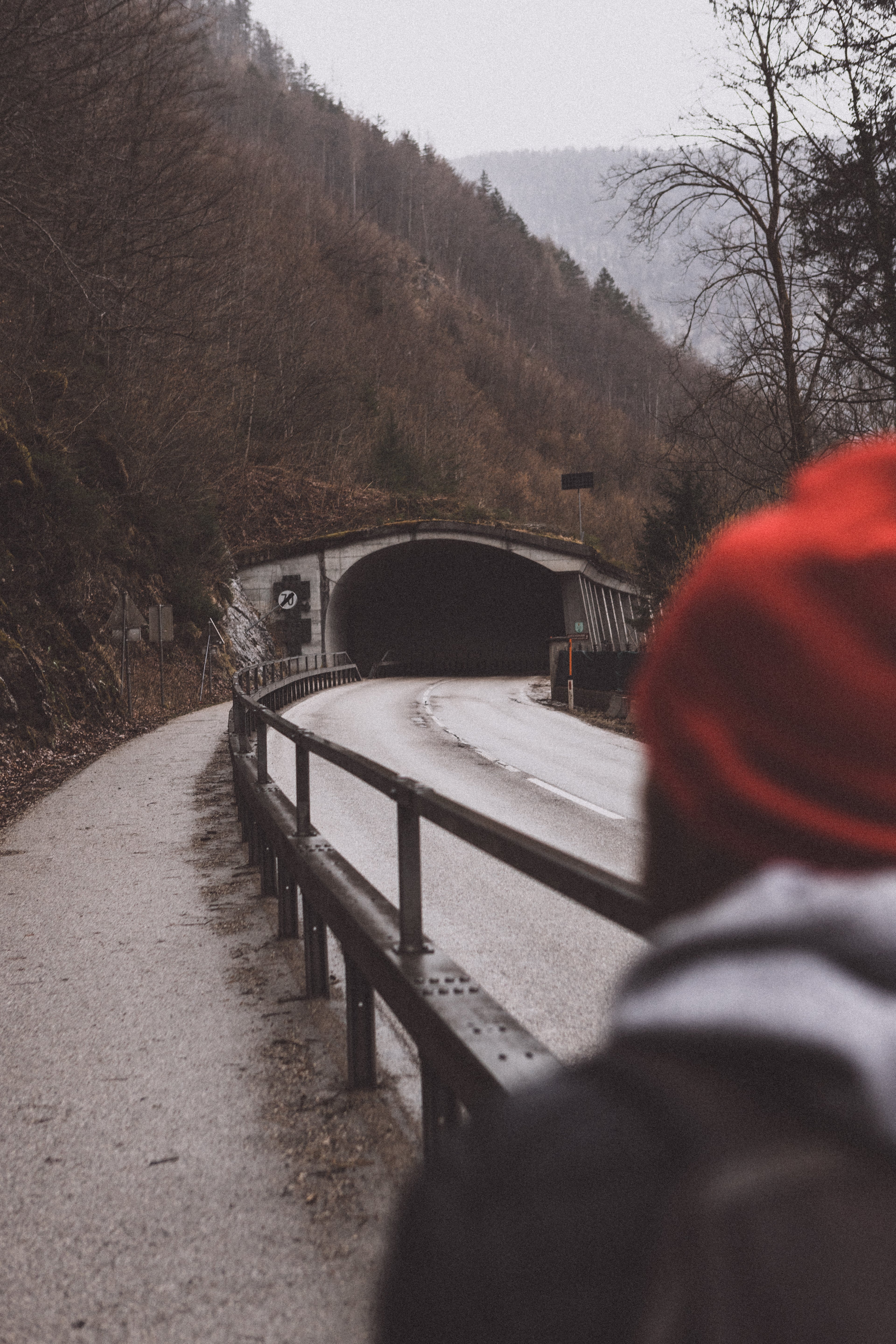 Perspective of a traveler with a vibrant red beanie in the foreground, looking at a winding road leading into a mountain tunnel in Hallstatt.