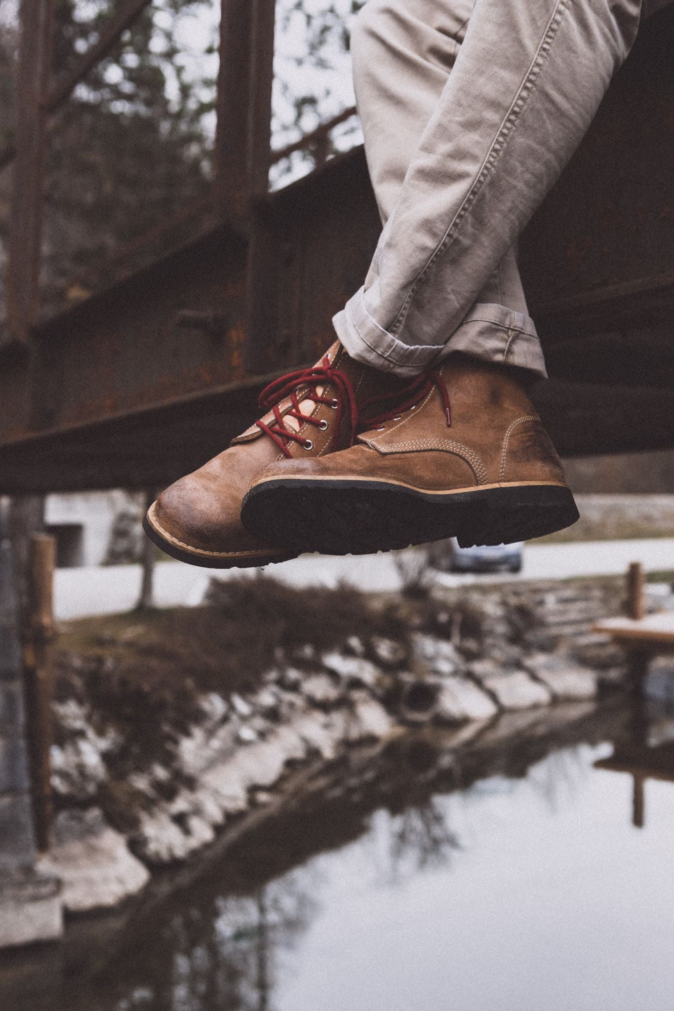 Person sitting on an elevated structure with legs dangling, wearing weathered leather Lobo Raw Boots with red laces in a mountain setting.