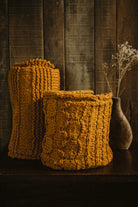 Two mustard yellow Re-Born crochet scarves, handmade from repurposed materials, displayed in a rustic setting next to a vase with dried flowers.