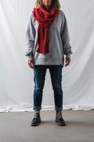 Woman wearing a bright red handmade knit Re-Born Scarf paired with a grey sweatshirt and jeans in a studio setting.