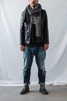 Man in an urban look wearing a grey knit Re-Born Scarf, black leather jacket, and jeans in a studio with a light background.