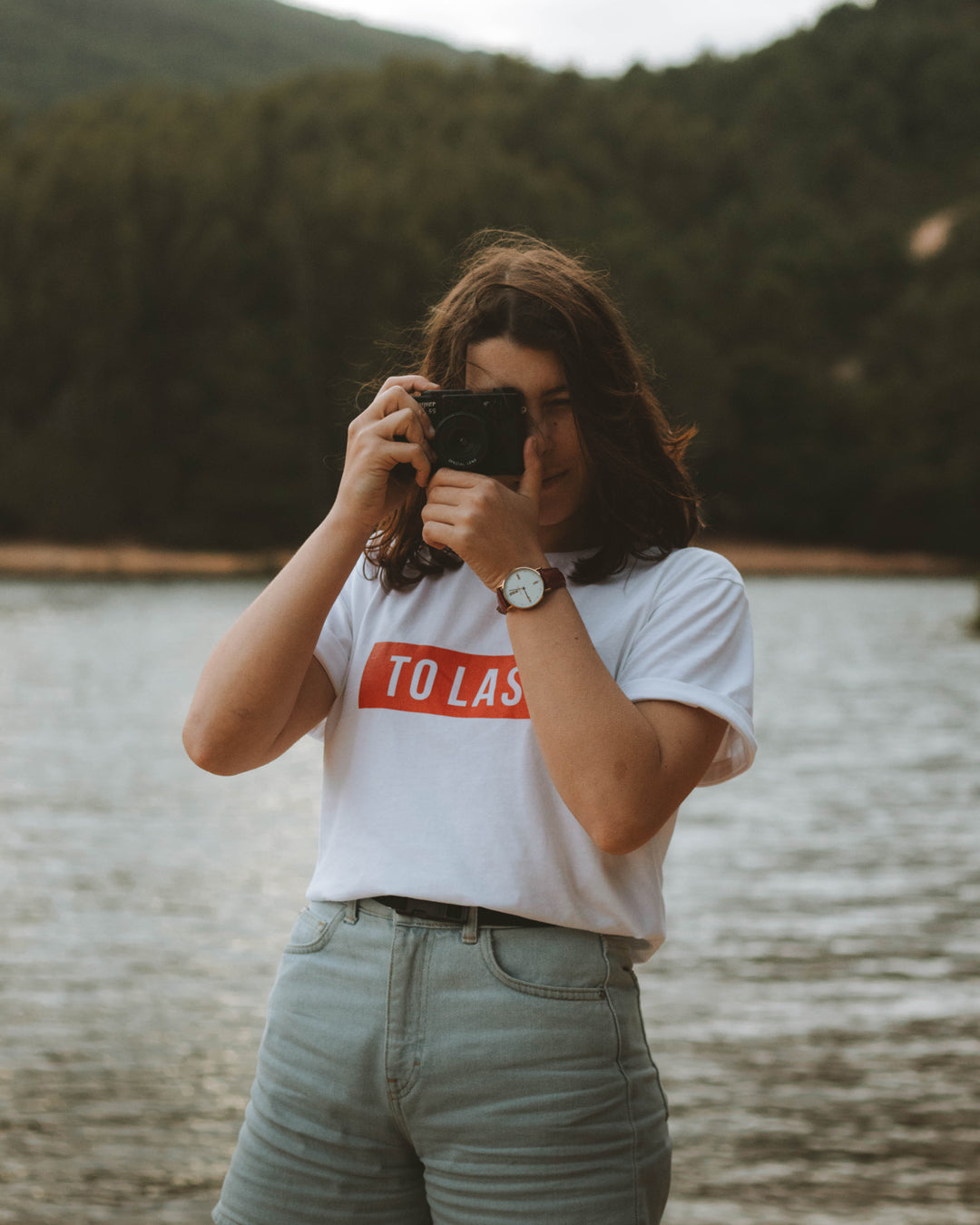 Close-up of a woman taking a photo with an analogue camera, wearing the white To Last t-shirt with a red logo.