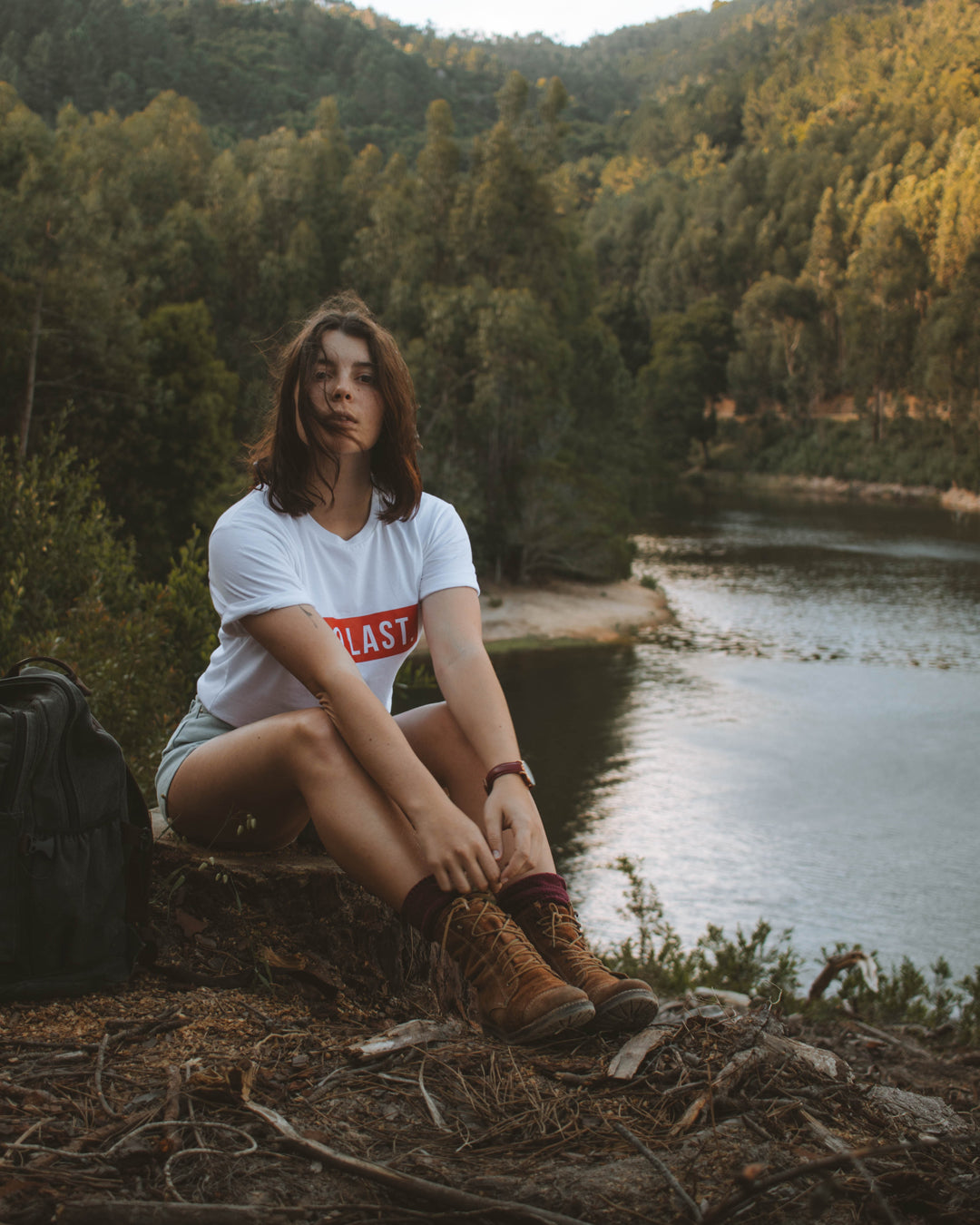 Woman sitting by the river wearing hiking boots, denim shorts, and the white To Last t-shirt.